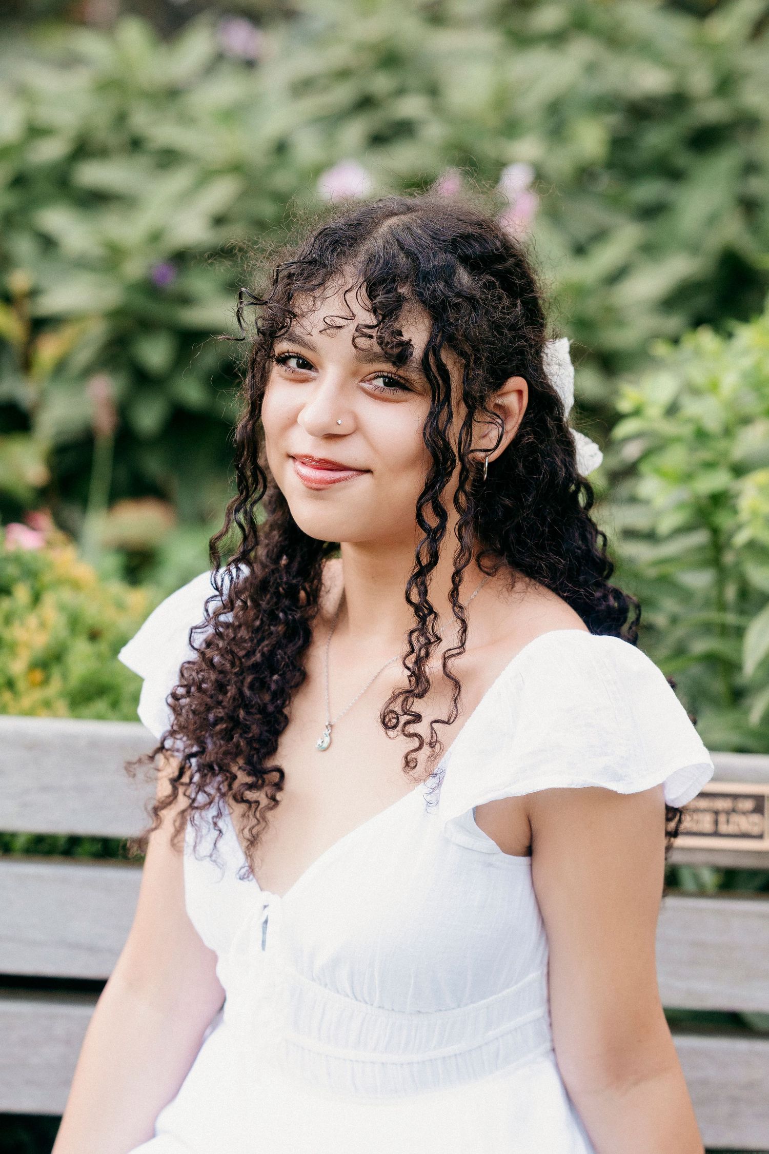 A person in a white ruffled dress sits on a wooden bench surrounded by lush greenery on a sunny day.