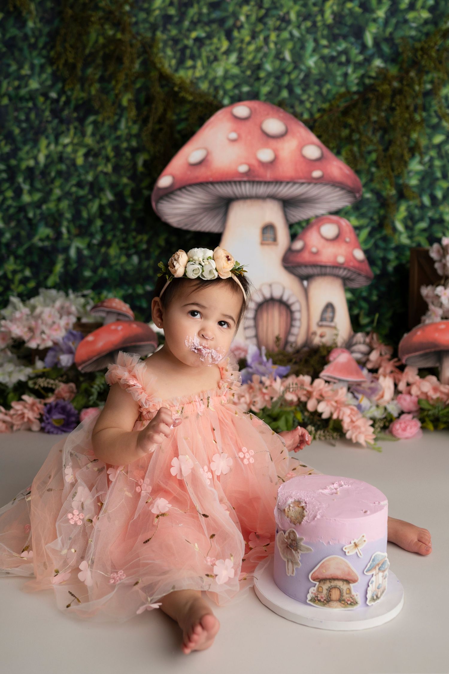 Enchanting fairy garden cake smash setup with pink mushroom houses, flowers, and a delicate purple and pink birthday cake.