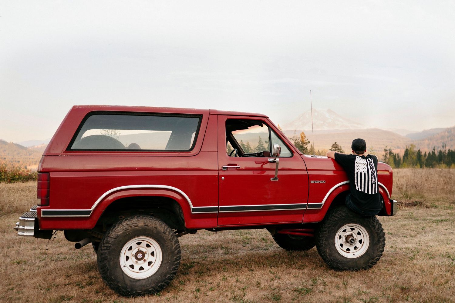 A red vintage Ford Bronco SUV parked in a desert landscape with mountains in the background.