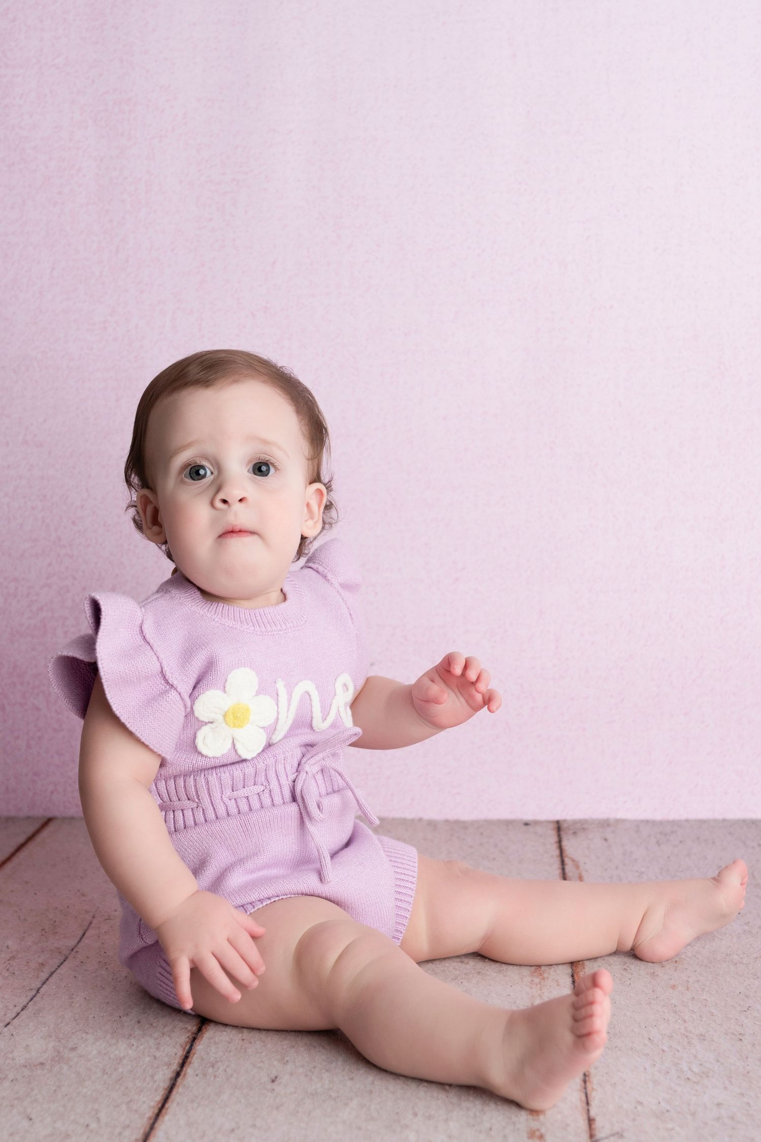 A baby girl in a purple “one” romper sits barefoot on a wooden floor against a soft pink background.