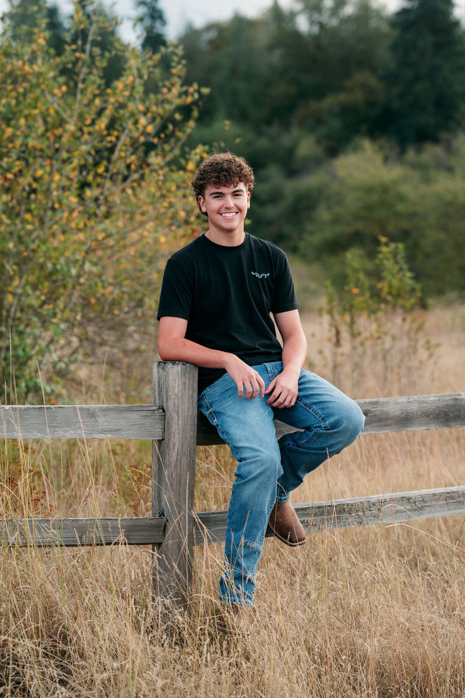 Young man with curly hair smiling, seated on a wooden fence in jeans and black tee, surrounded by nature.