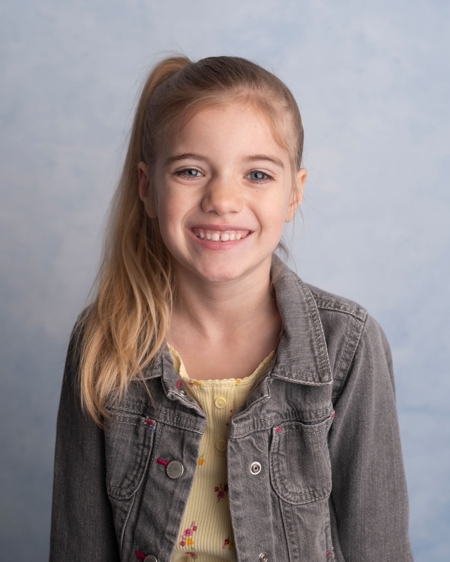 Young student wearing a gray denim jacket and yellow top poses for a portrait against a mottled blue background.