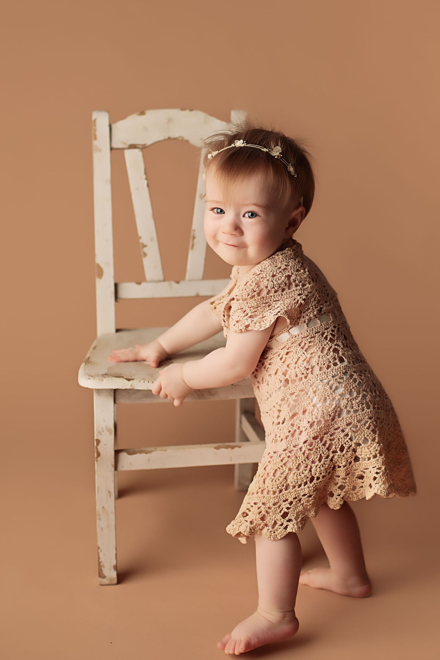 Small child in lace dress stands against brown background holding white vintage chair.