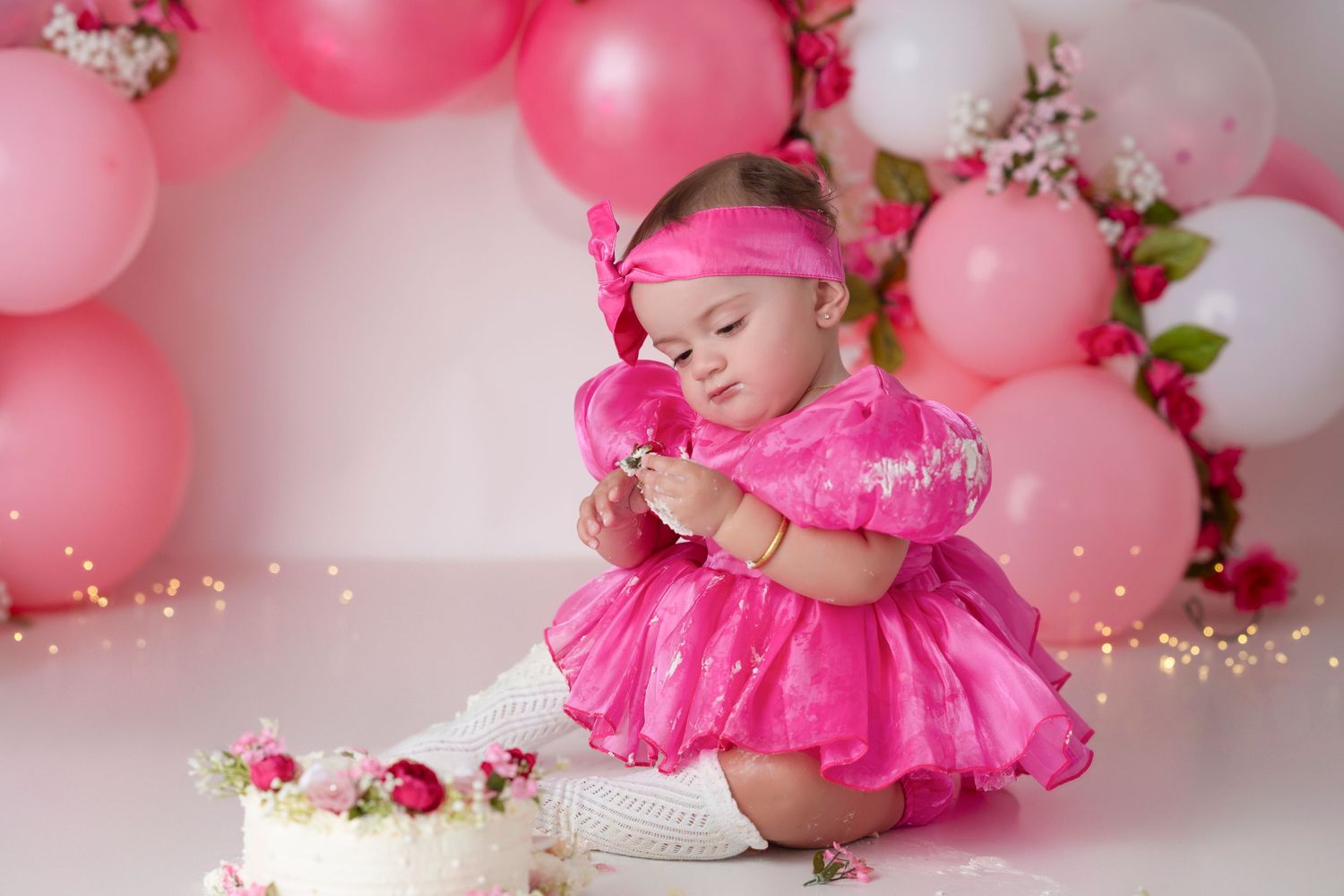 A baby wearing a pink tutu dress poses for milestone photos with pink and white balloons and floral decorations.