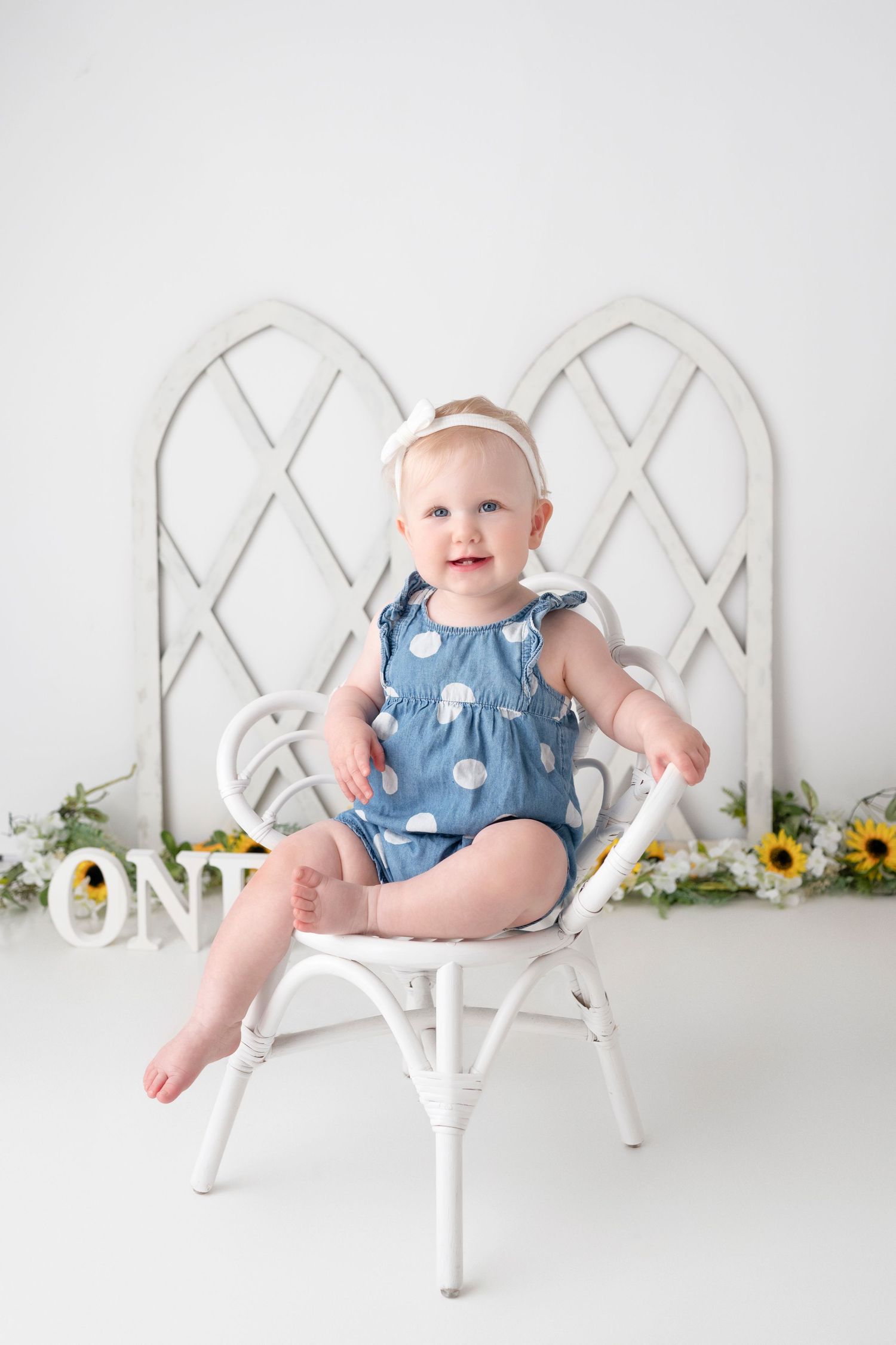 A baby in a polka dot dress sits on a white lattice chair with sunflowers in the background.