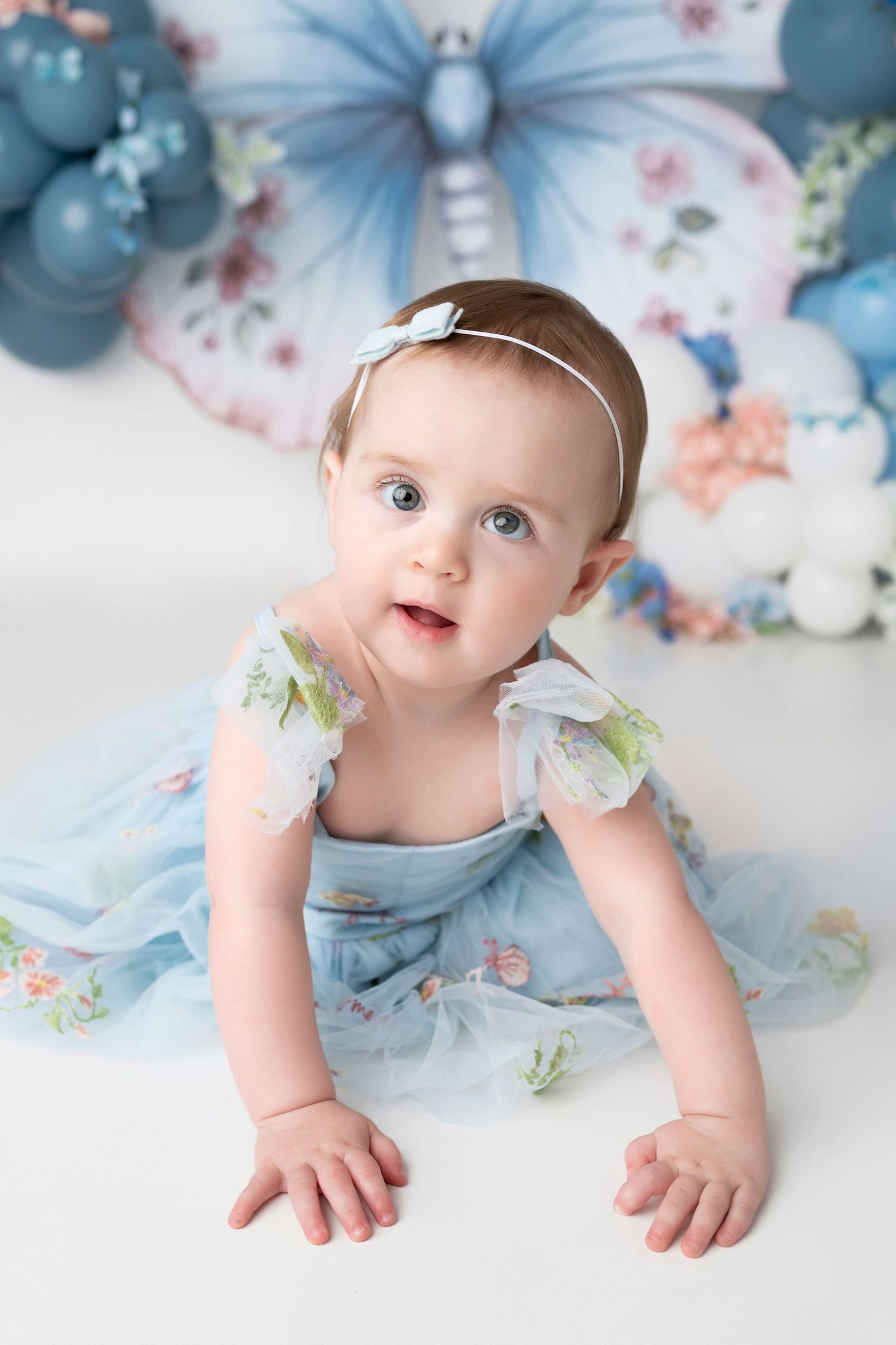 Sweet portrait with a blue floral dress against a pastel butterfly and balloon backdrop.