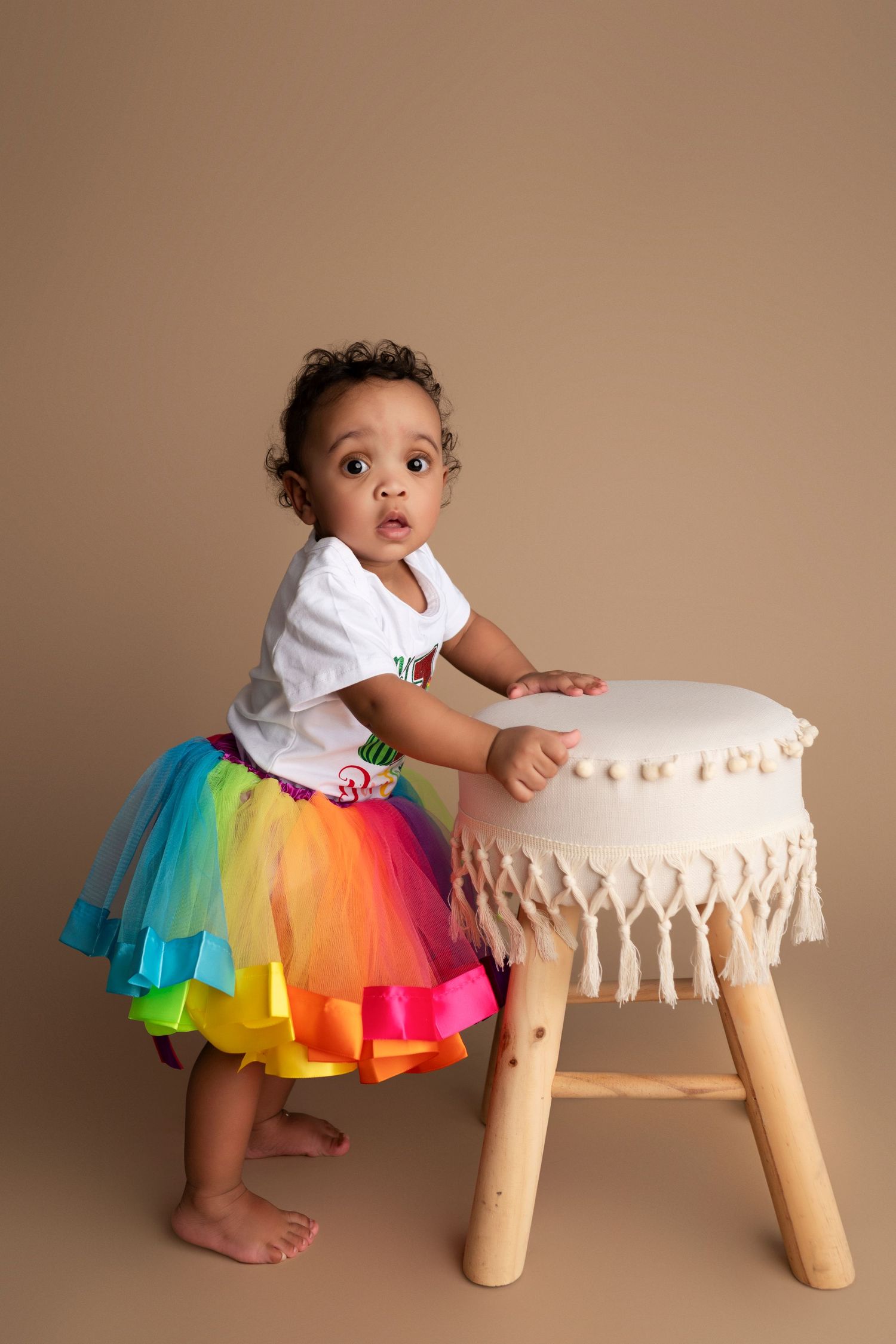 A toddler in a vibrant rainbow tutu skirt stands beside a white cushioned stool against brown backdrop.