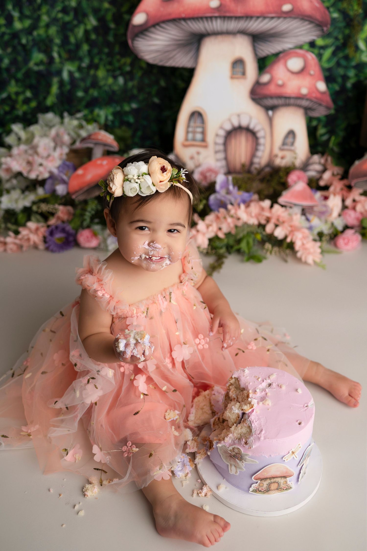A cake smash photo session featuring pink decor, mushroom props, and a white cake against a floral backdrop.