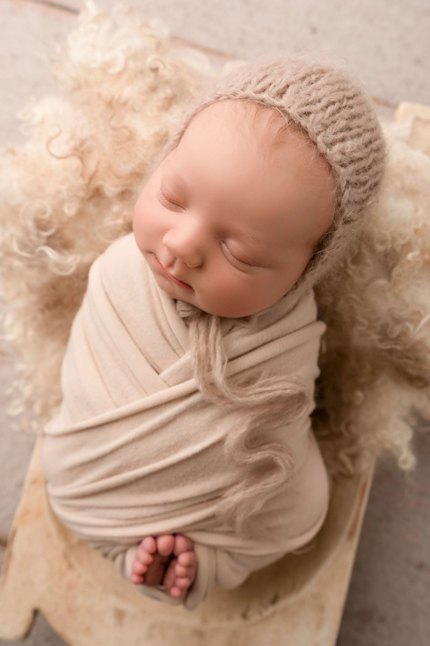 A sleeping newborn baby swaddled in soft cream fabric and wearing a knit bonnet rests on a textured backdrop.