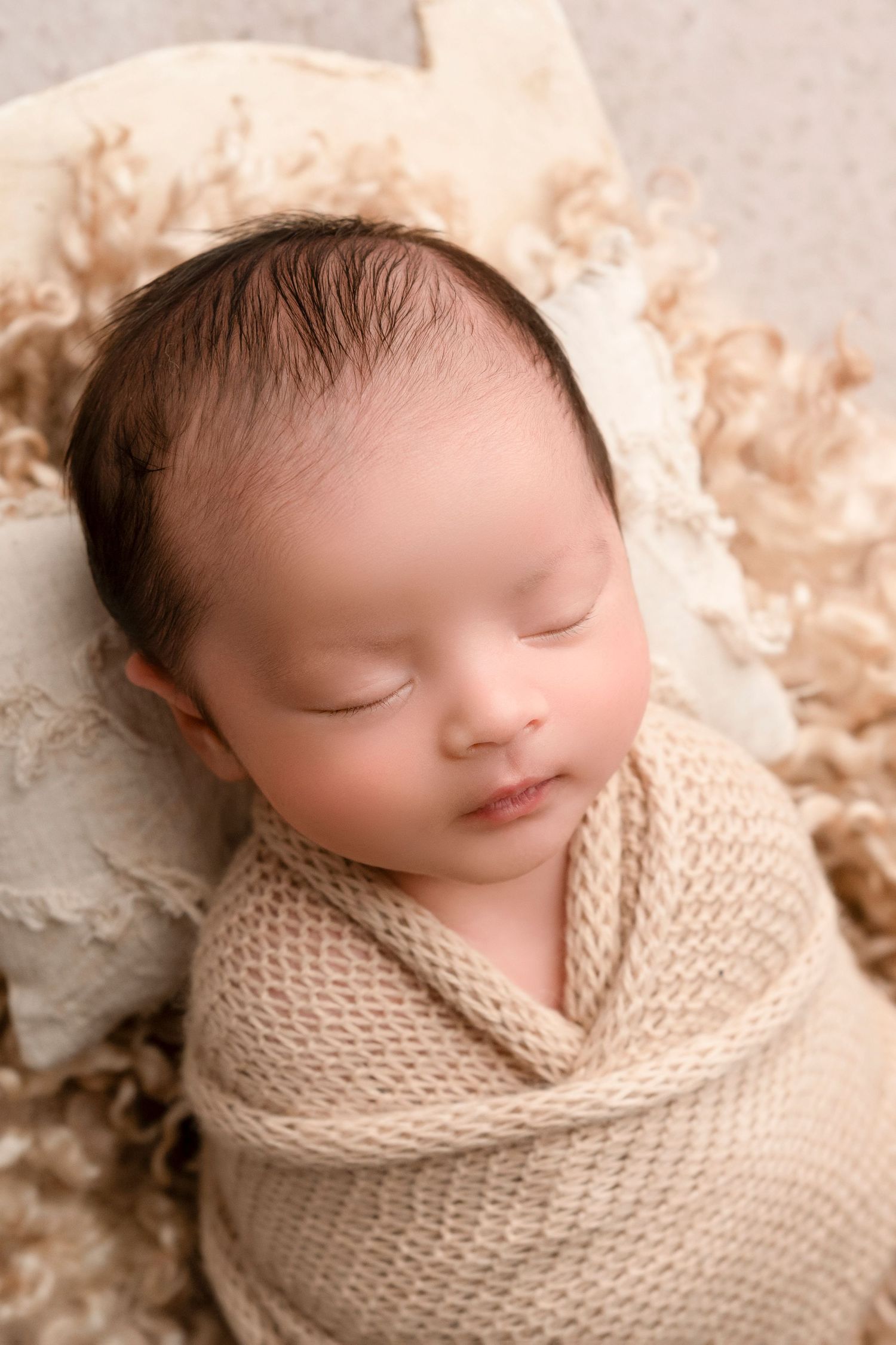 Peaceful newborn swaddled in soft knitted wrap against neutral cream background.