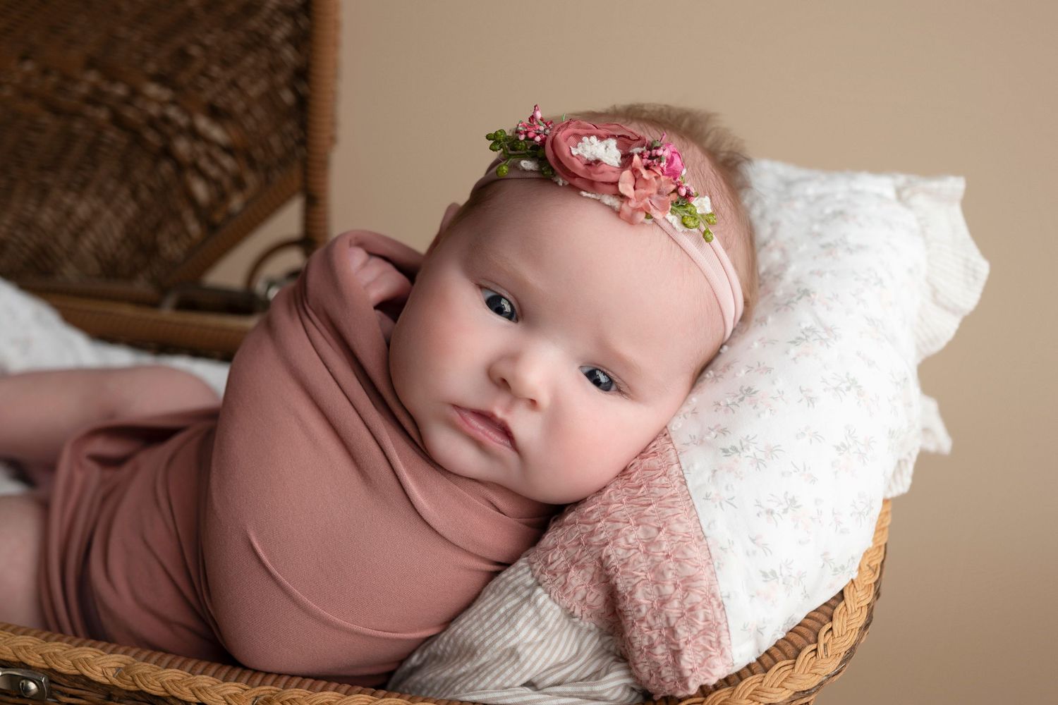 Newborn baby girl wrapped in dusty rose swaddle wearing floral headband, posed in wicker basket.