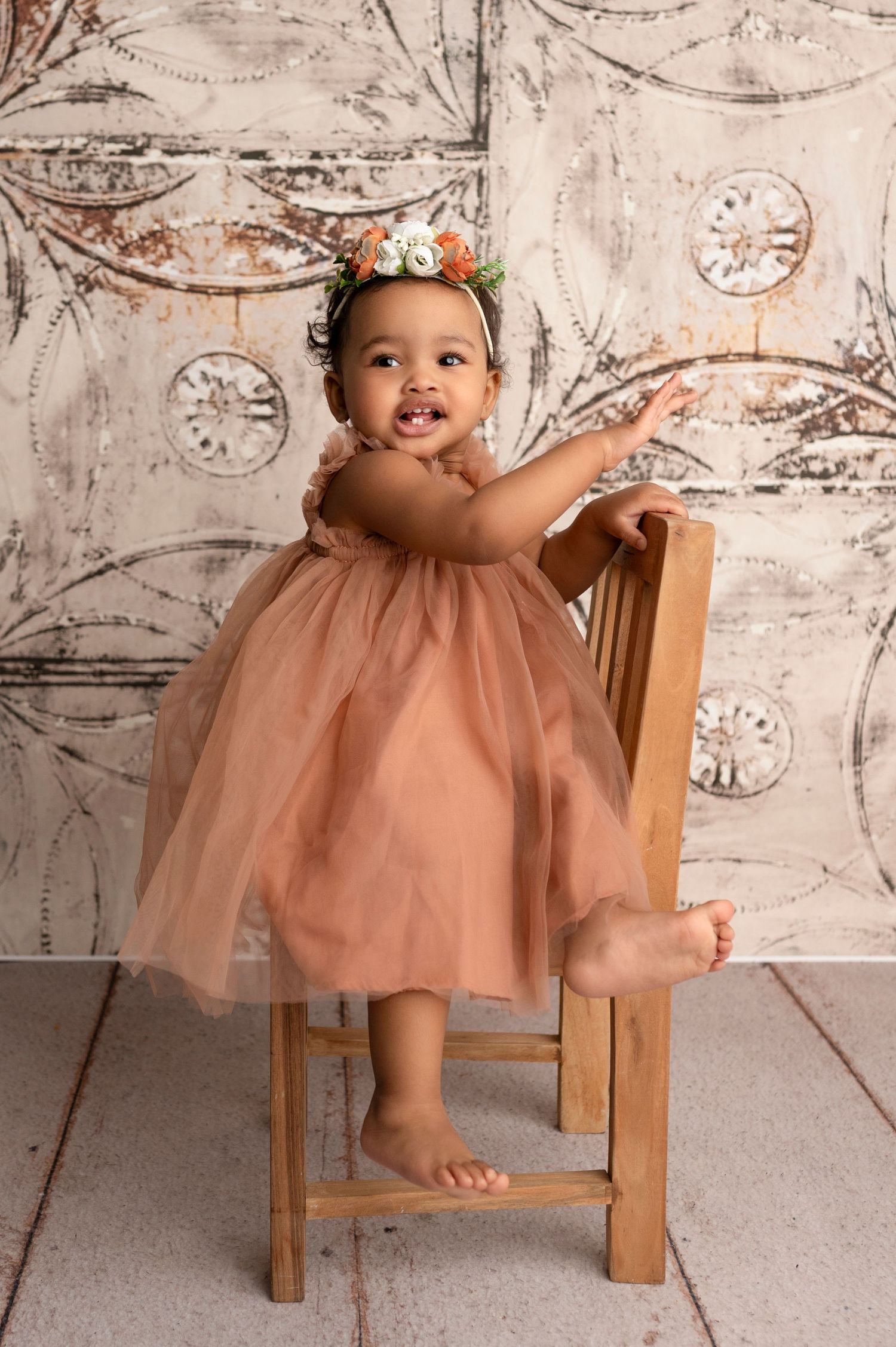 Smiling baby girl in a dusty rose tulle dress and floral crown standing by a wooden chair.
