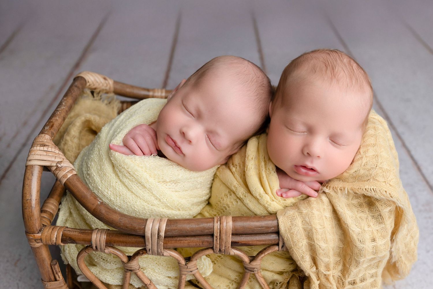 Two sleeping newborn twins wrapped in yellow swaddles nestled together in a wicker basket on a gray wood floor.