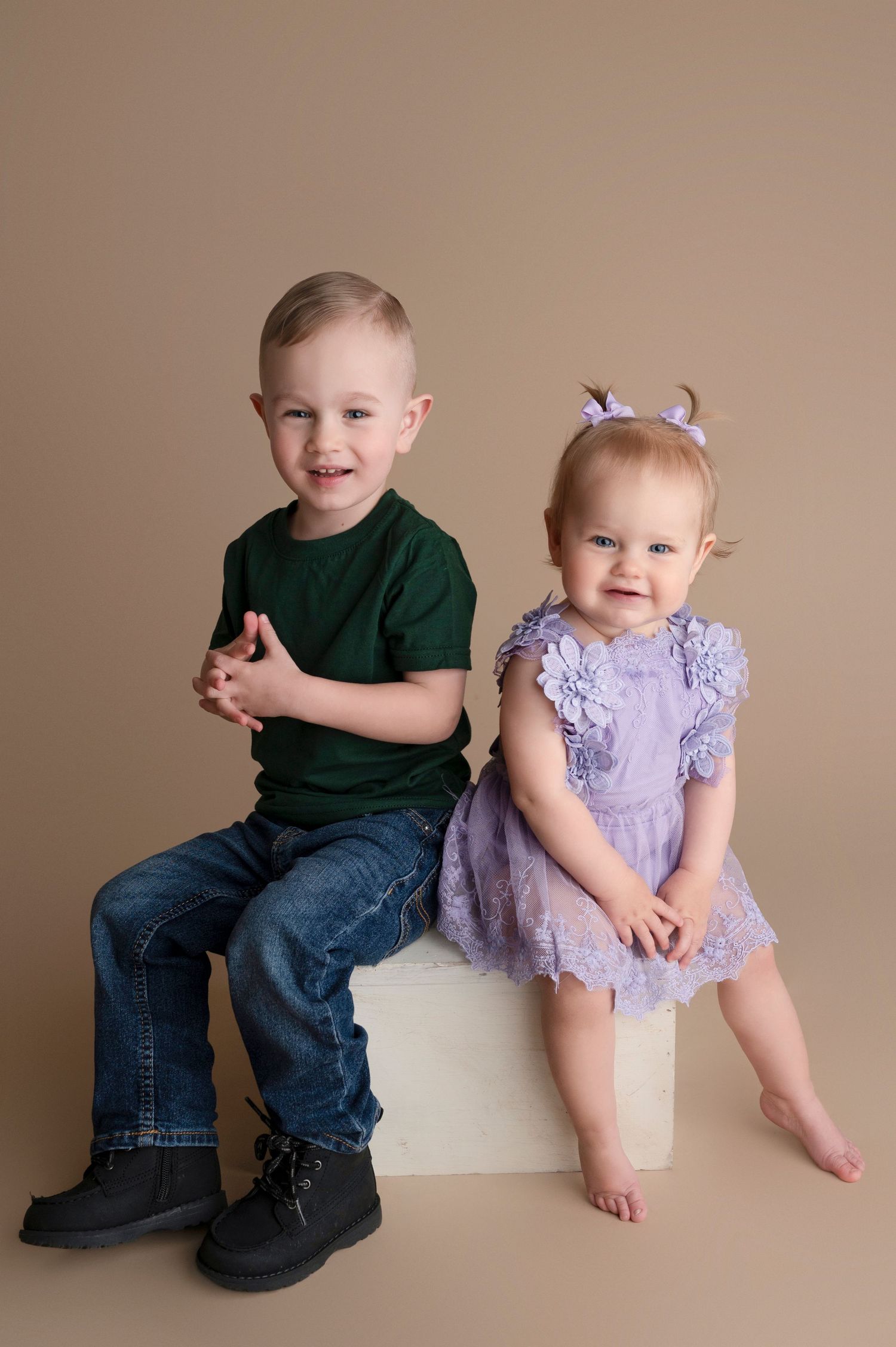Two young siblings pose for a studio portrait, boy in green shirt and jeans, baby girl in purple dress on white cube.