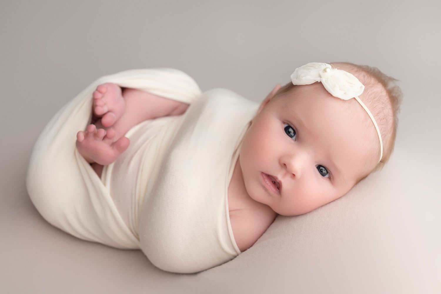 Newborn baby girl swaddled in white wrap with bow headband, posed on gray background.