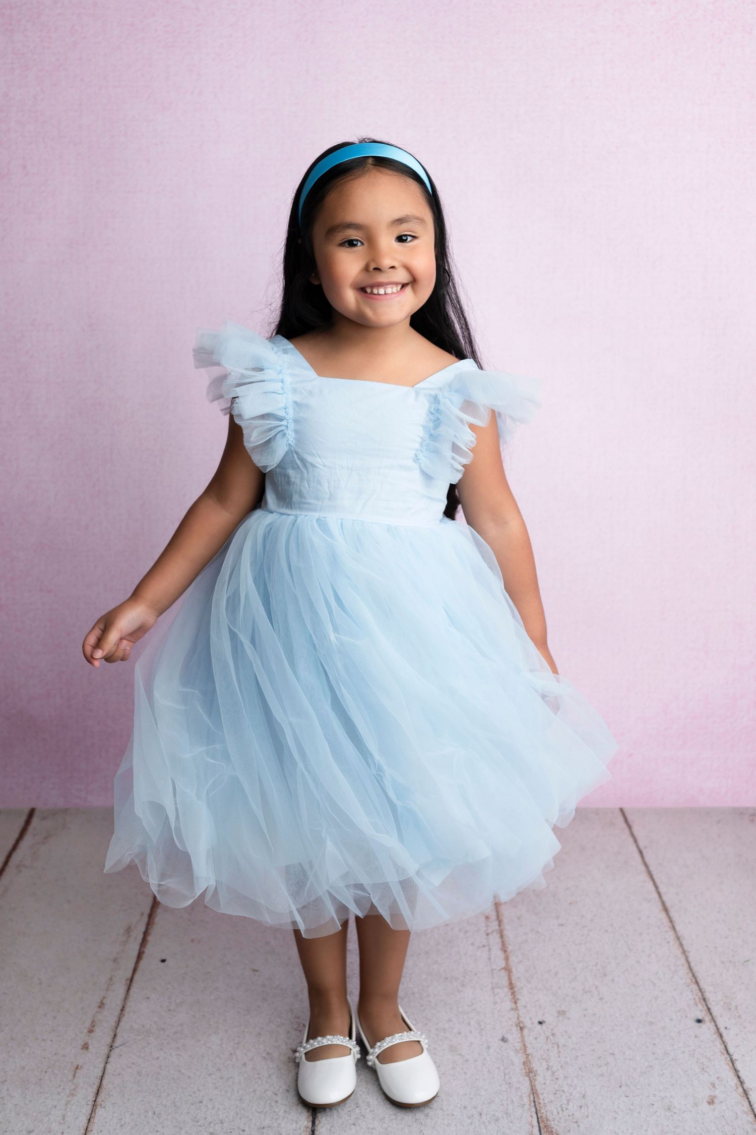 Young girl smiling in a light blue tulle dress with ruffle sleeves and headband, posing against a pink background.