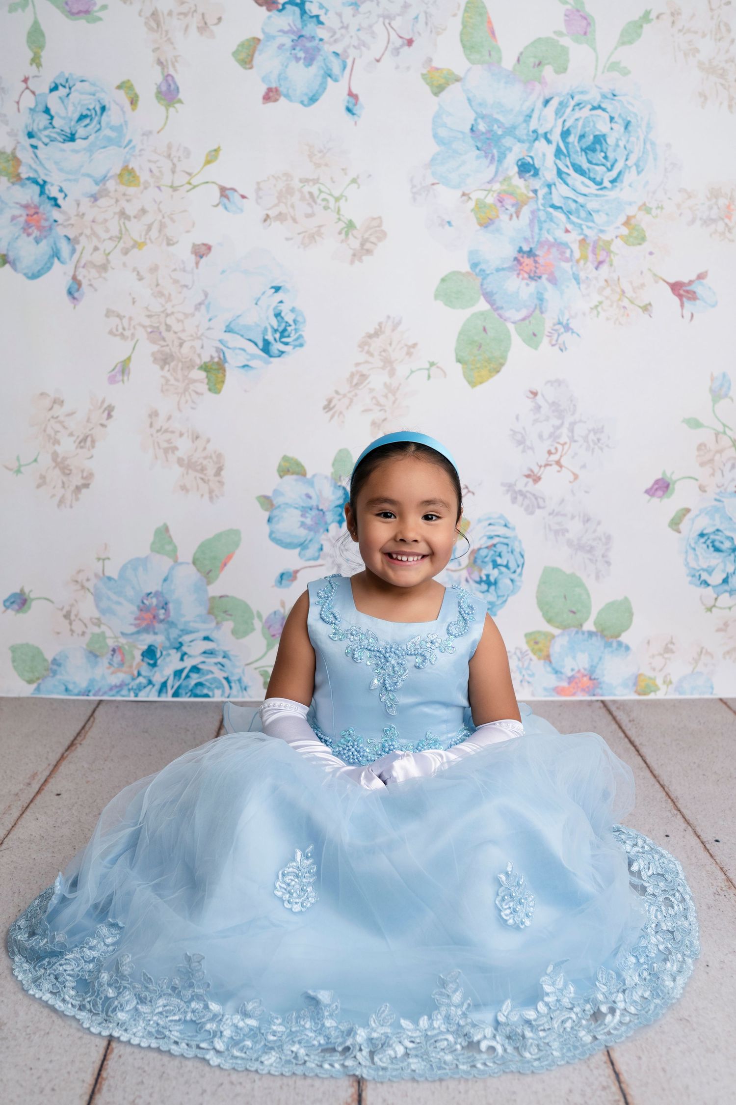 Young girl in blue princess gown and gloves smiles against floral backdrop.