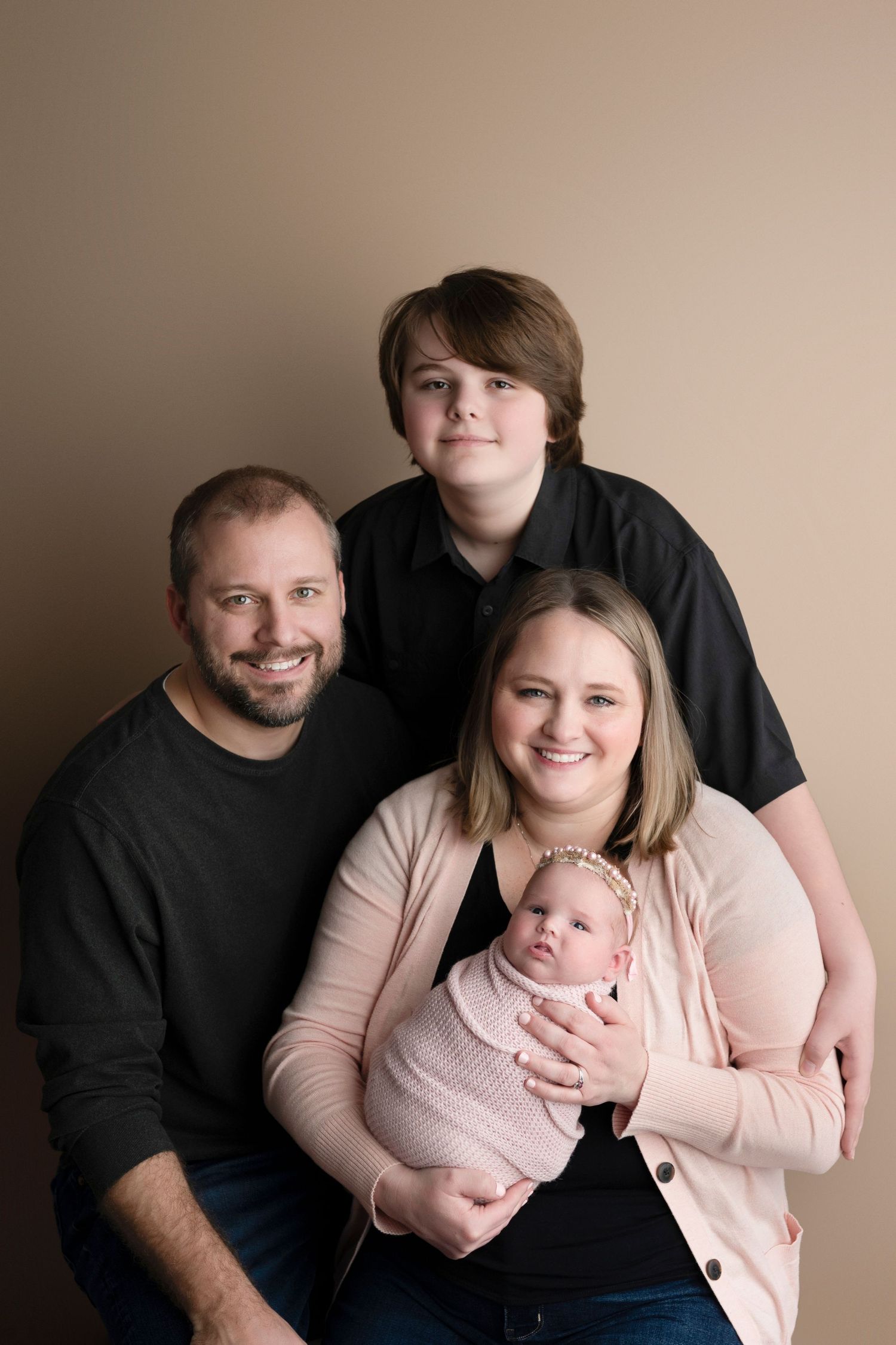 Family portrait with parents, teen son, and newborn baby girl in pink wrap, posed against a warm beige background.