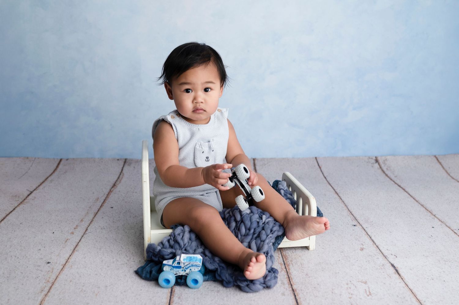 A baby boy sits in a small wooden bed with gray knit blanket, holding toy cars, against a soft blue background.