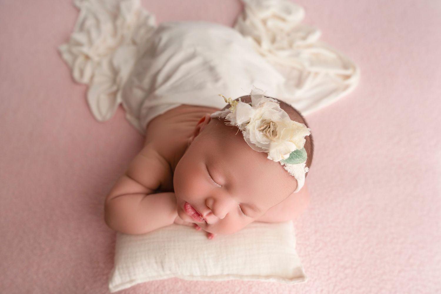 A sleeping newborn baby wearing a delicate white floral headband rests peacefully on pink fabric.