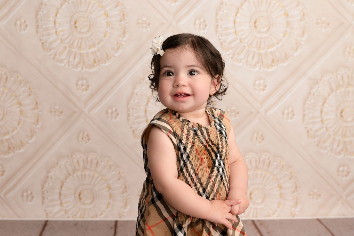 A toddler in a plaid dress sits against a cream-colored textured backdrop for a photo session.