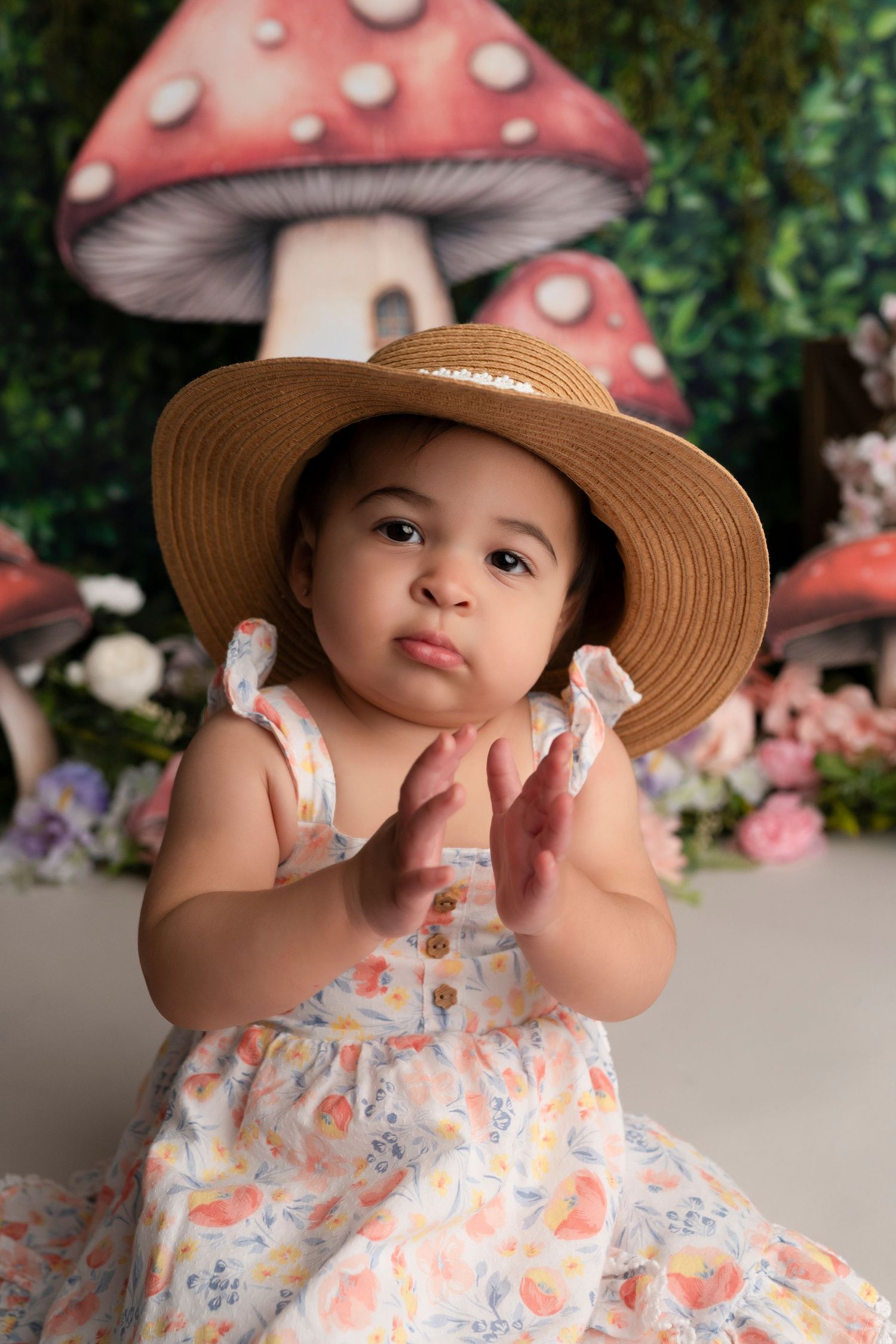 A baby in a floral dress and straw hat poses in front of a whimsical mushroom backdrop with pink flowers.