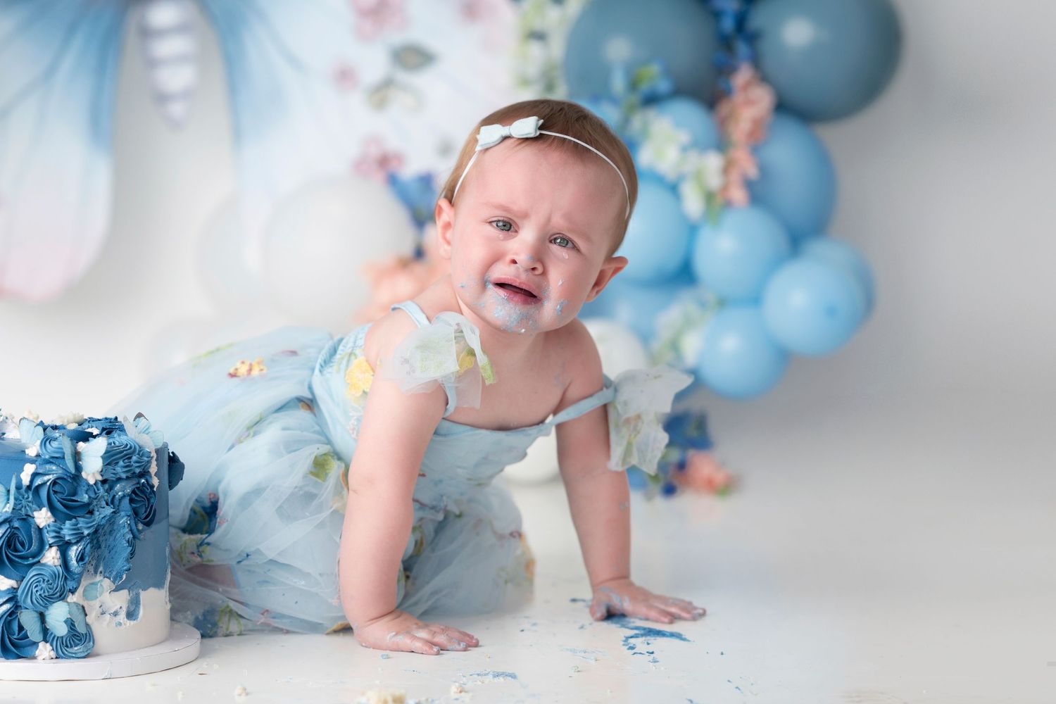 Baby crawling on white floor with blue balloon garland and butterfly decorations in dreamy studio setting.