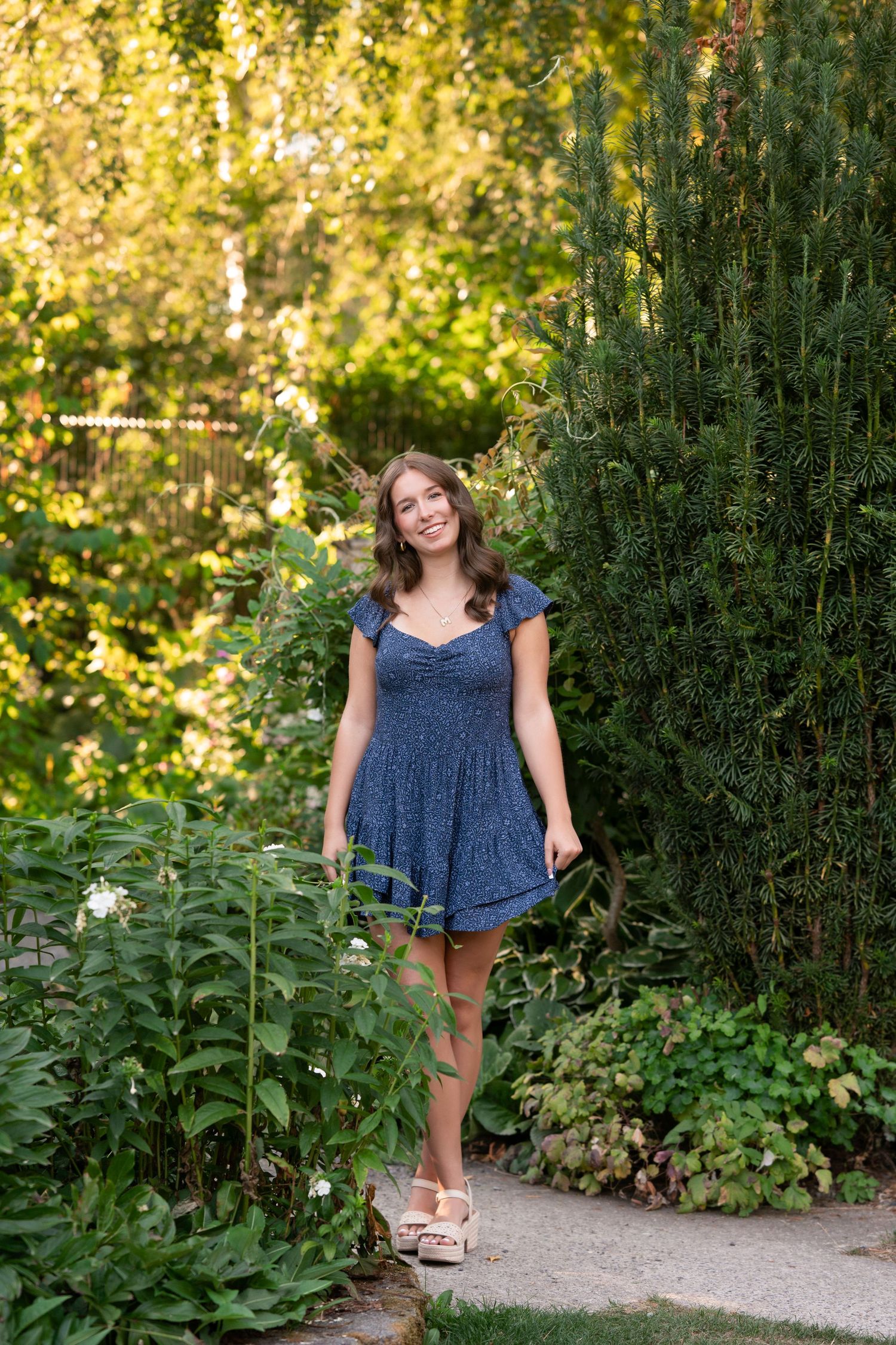 A person in a blue floral dress poses in a lush garden path surrounded by greenery on a sunny summer day.