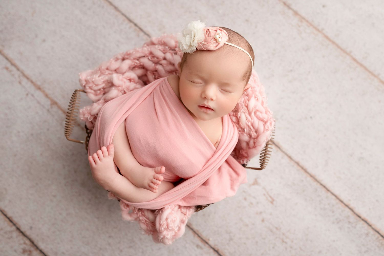 Newborn baby in pink wrap sleeping peacefully on white wooden floor during professional photo session.