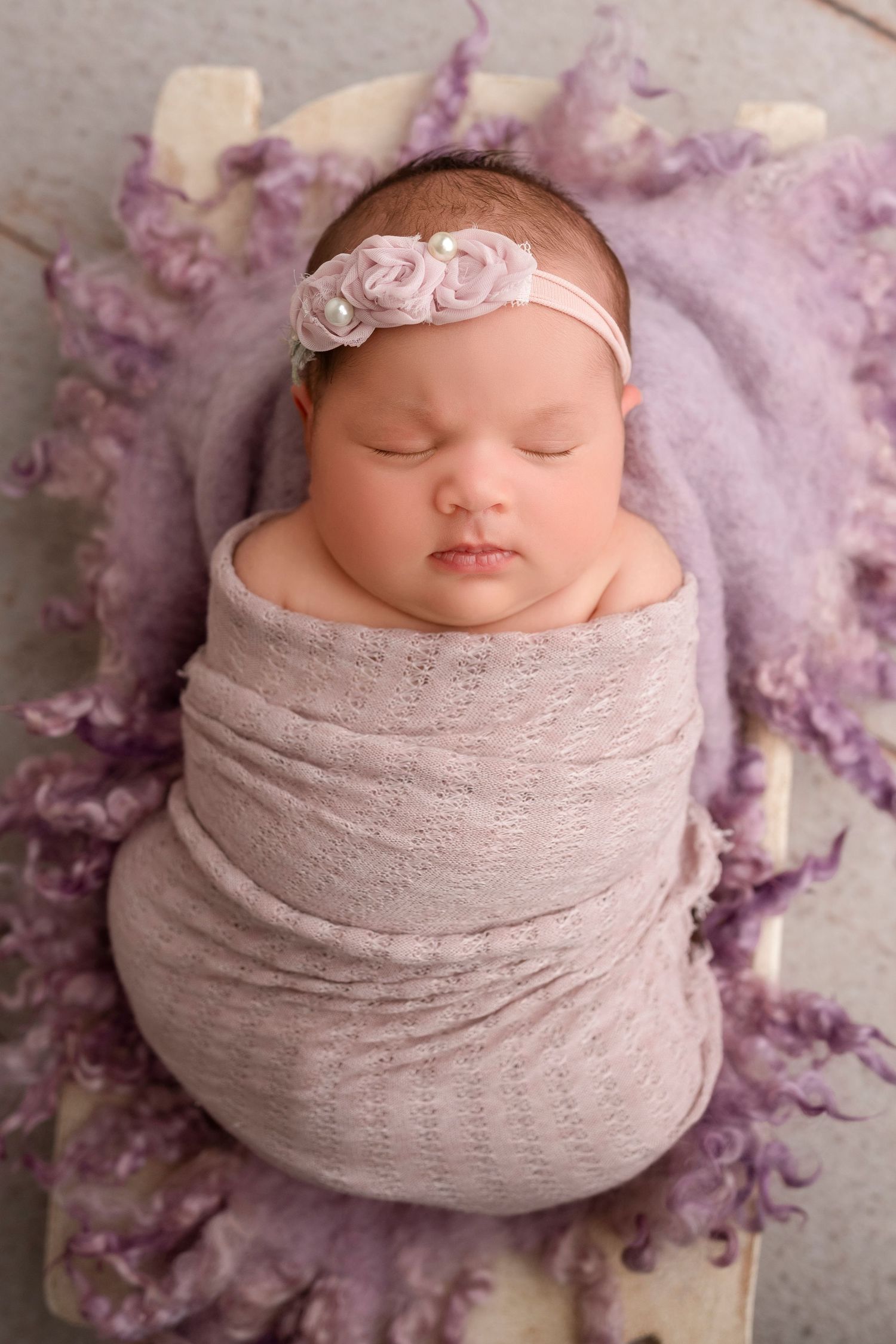 Sleeping newborn wrapped in soft gray swaddle with floral headband on purple textured backdrop.