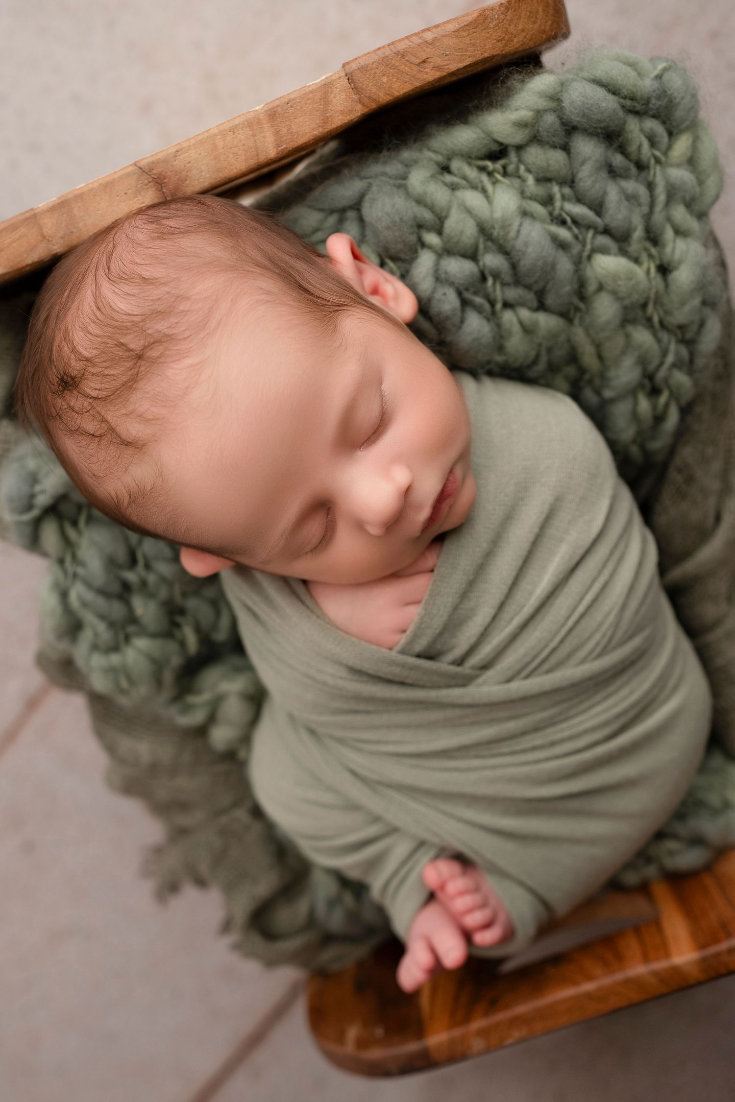 Sleeping baby wrapped in sage green swaddle lying on wooden surface with textured blanket.