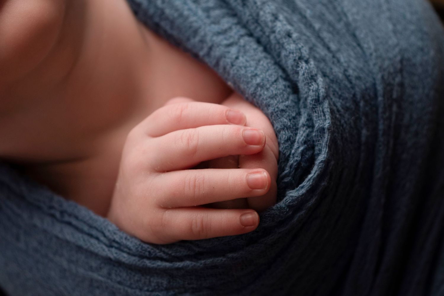 A tiny baby's hand rests gently on soft blue-gray fabric in a tender close-up shot.