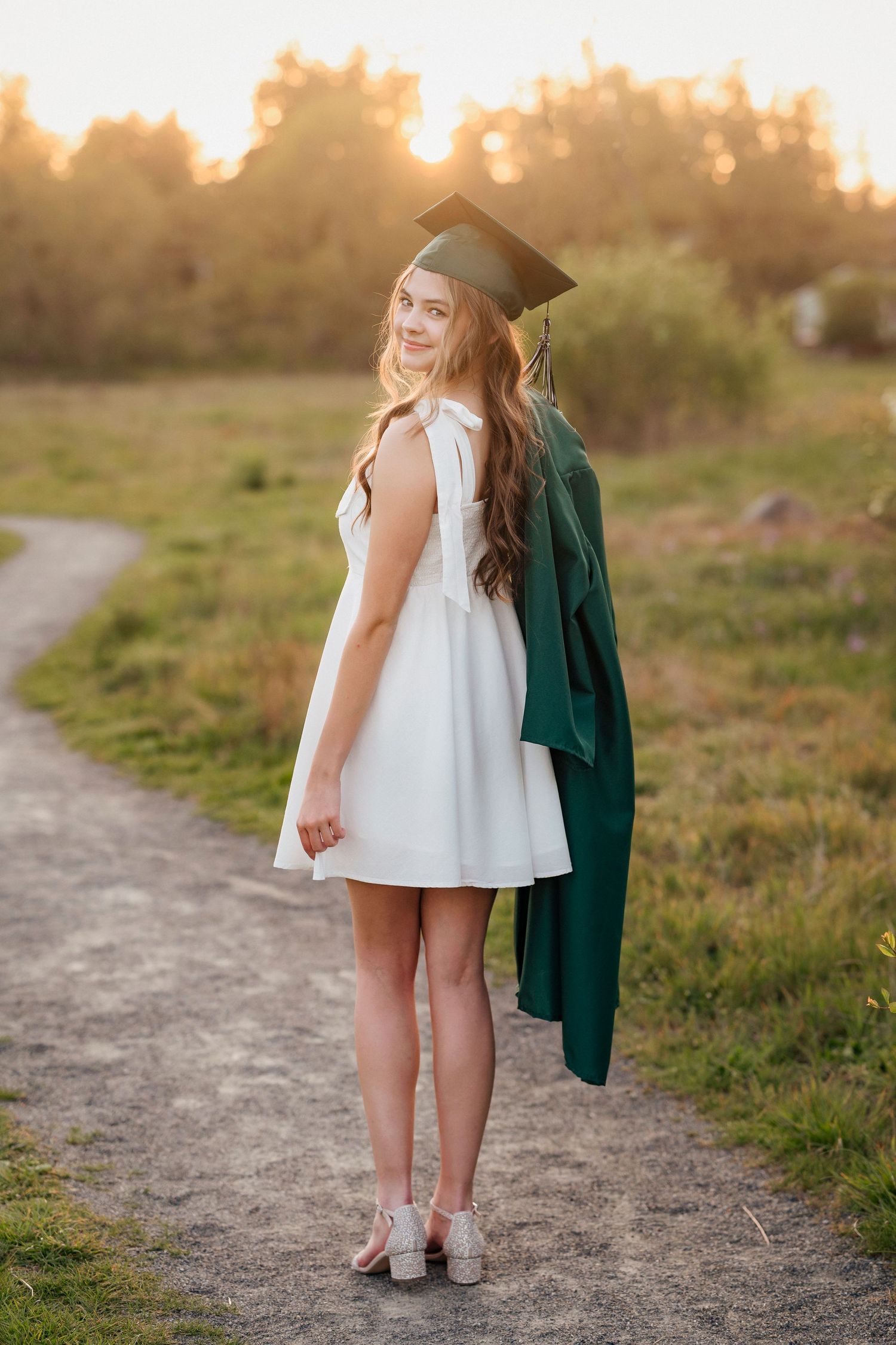Graduate in white dress and green gown stands on dirt path during golden sunset holding cap.