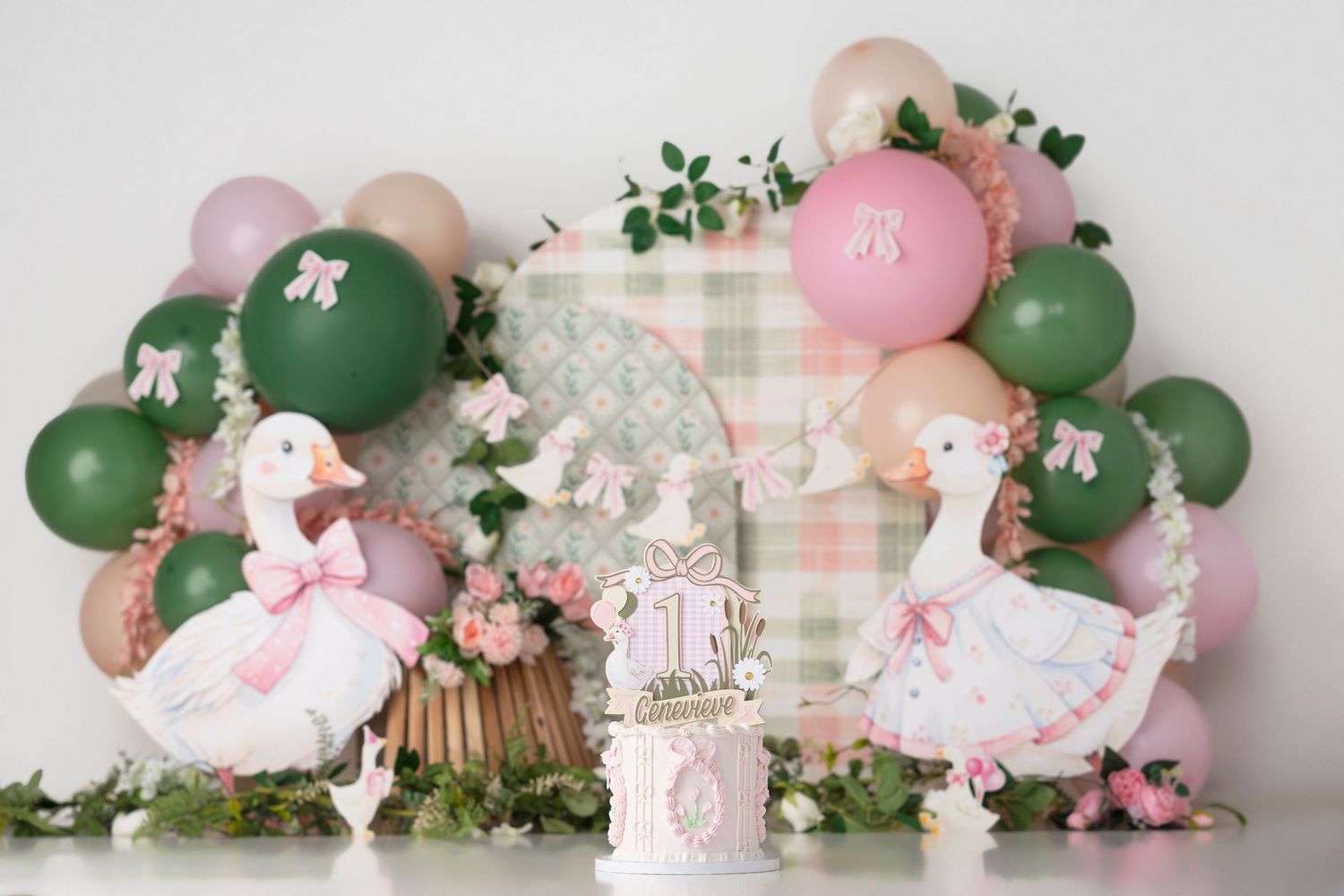 First birthday cake smash setup with white geese, pink and green balloons, floral decorations, and a plaid backdrop.
