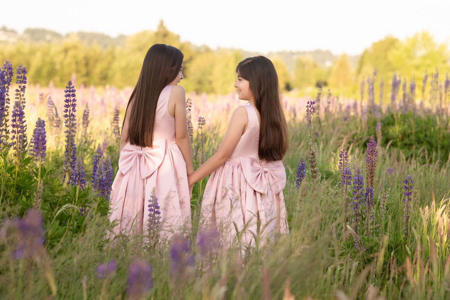 Two girls in matching pink dresses with bows hold hands in a blooming lupine field at golden hour.