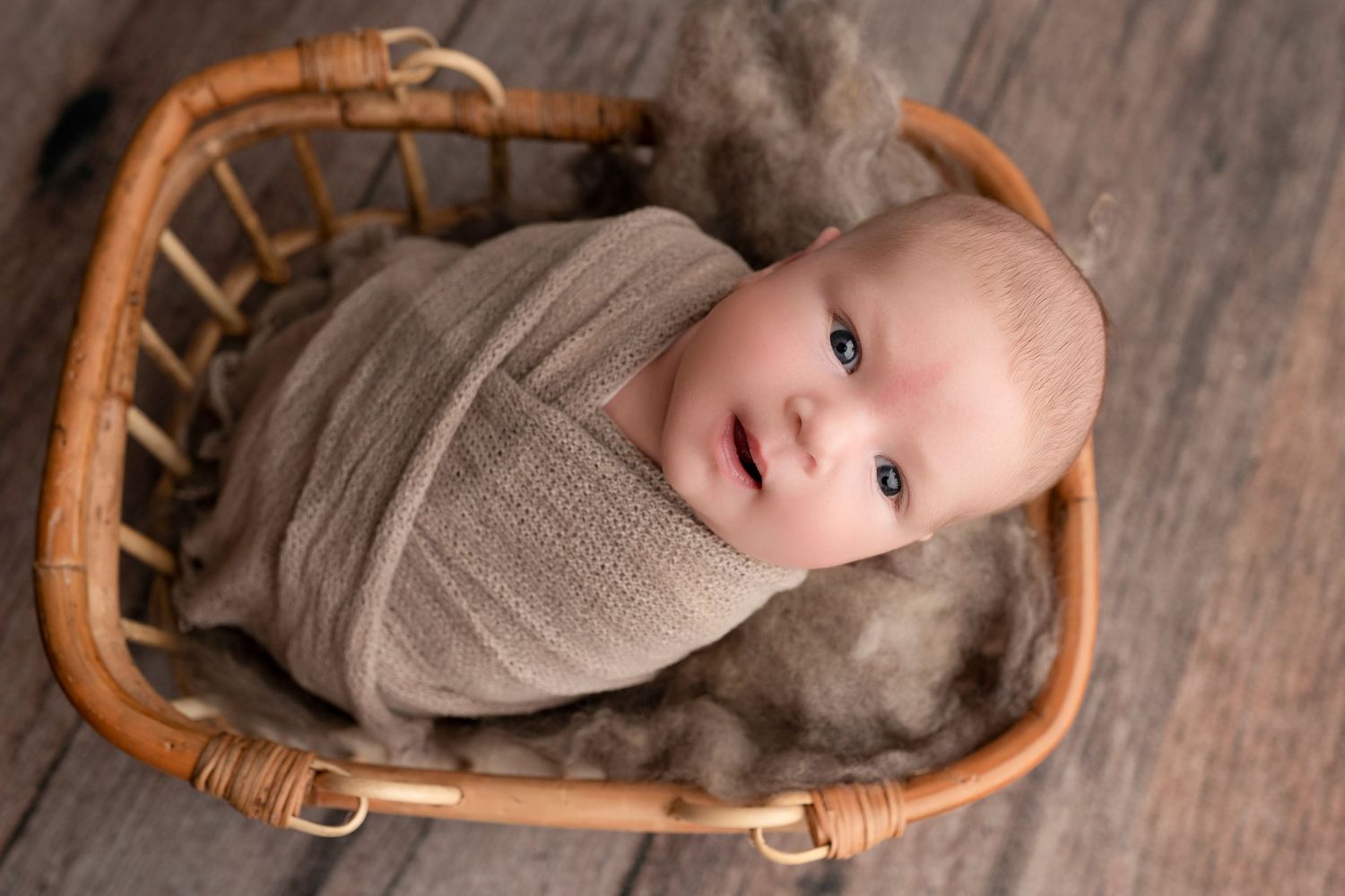 Newborn baby swaddled in a taupe wrap, nestled in a wicker basket with soft wool, gazing upward.