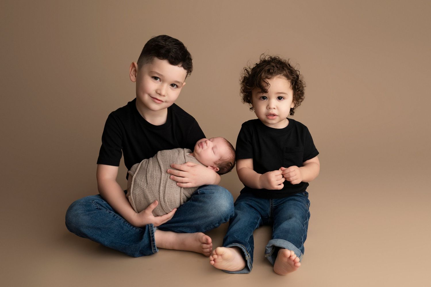 Two older brothers in black shirts and jeans sit together holding their newborn sibling on a tan studio backdrop.