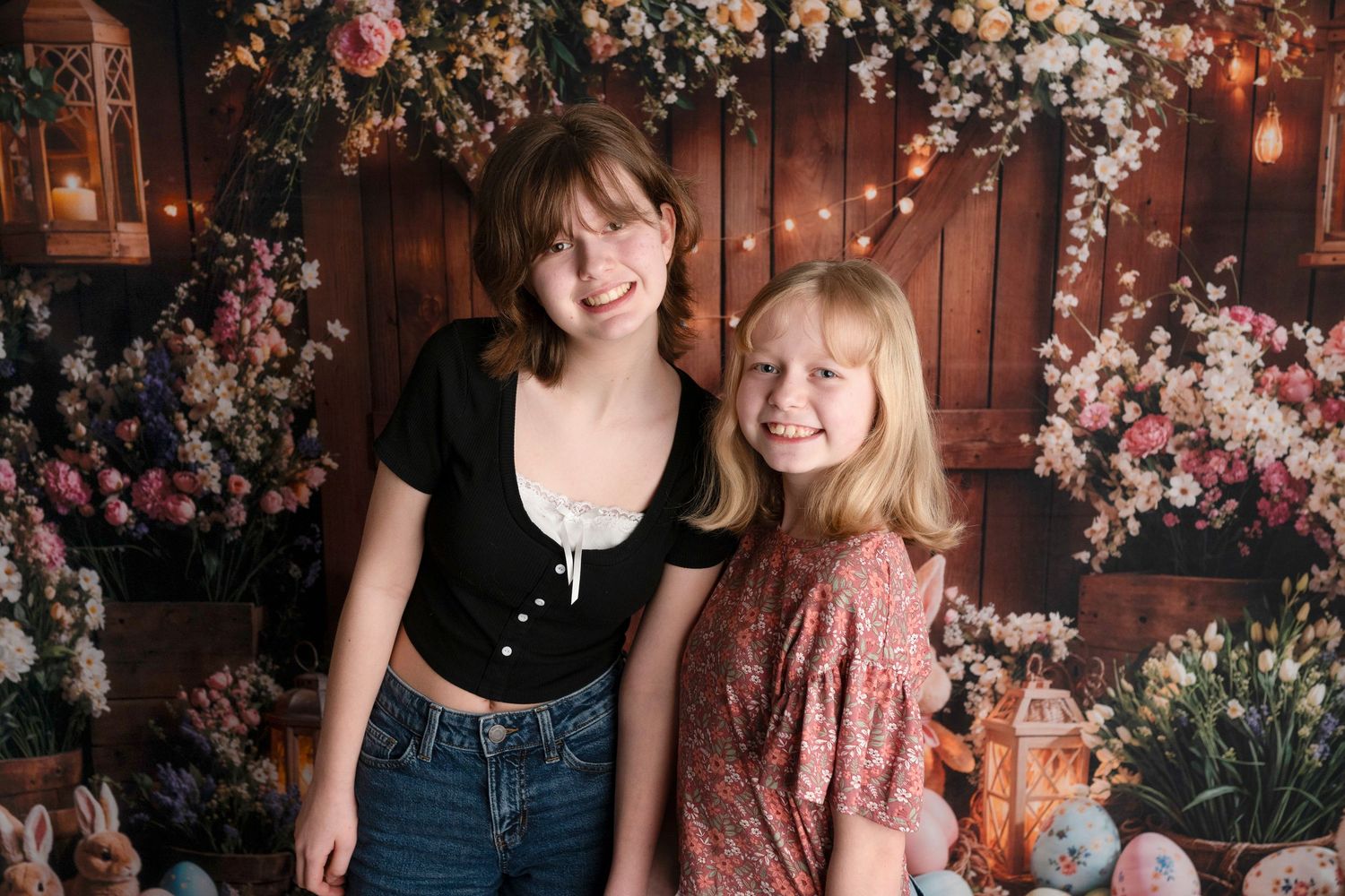 Two smiling girls pose together in front of a floral Easter-themed backdrop with soft lights and pastel decorations.