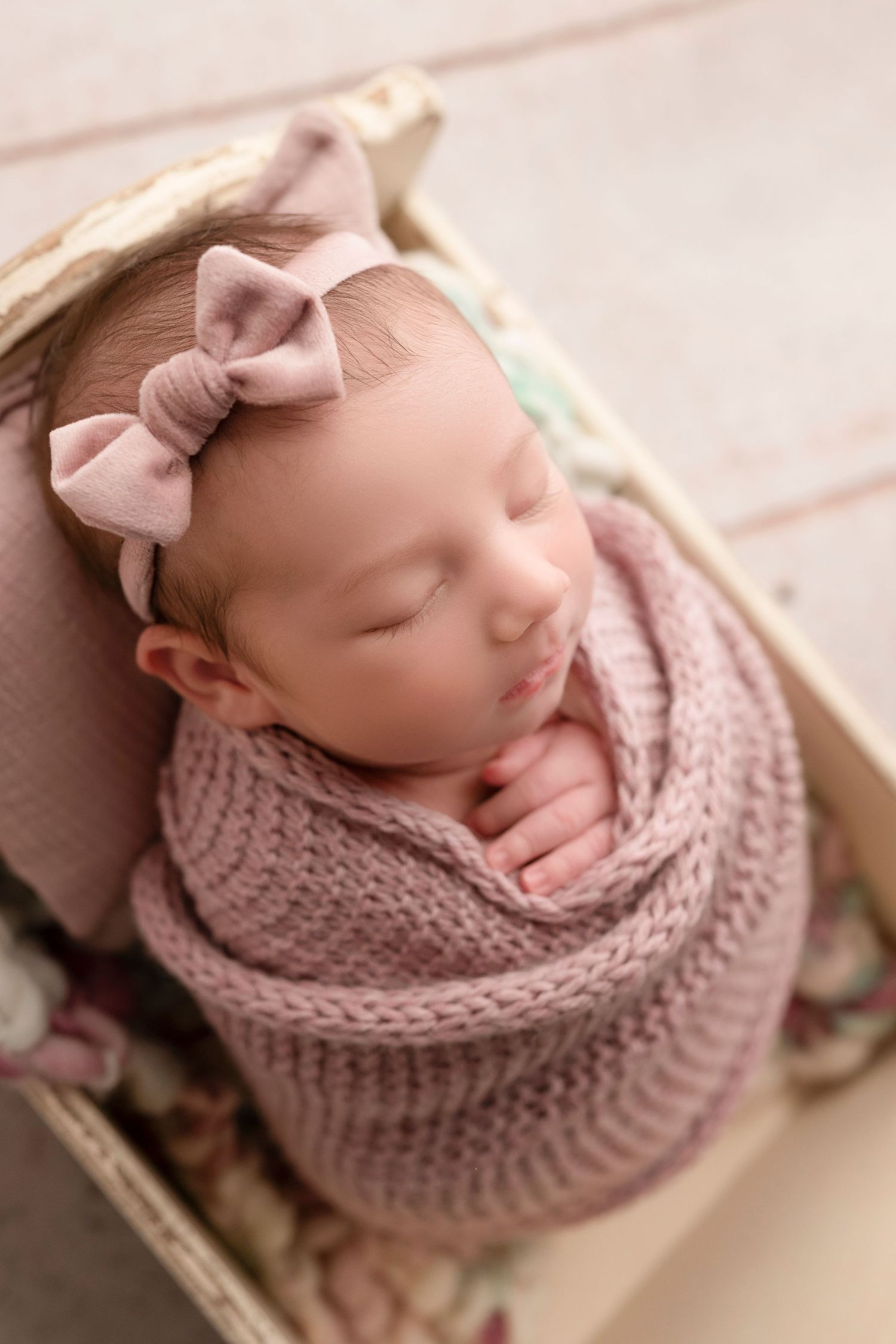 Sleeping newborn baby girl swaddled in pink knit wrap with bow headband in a wicker basket.