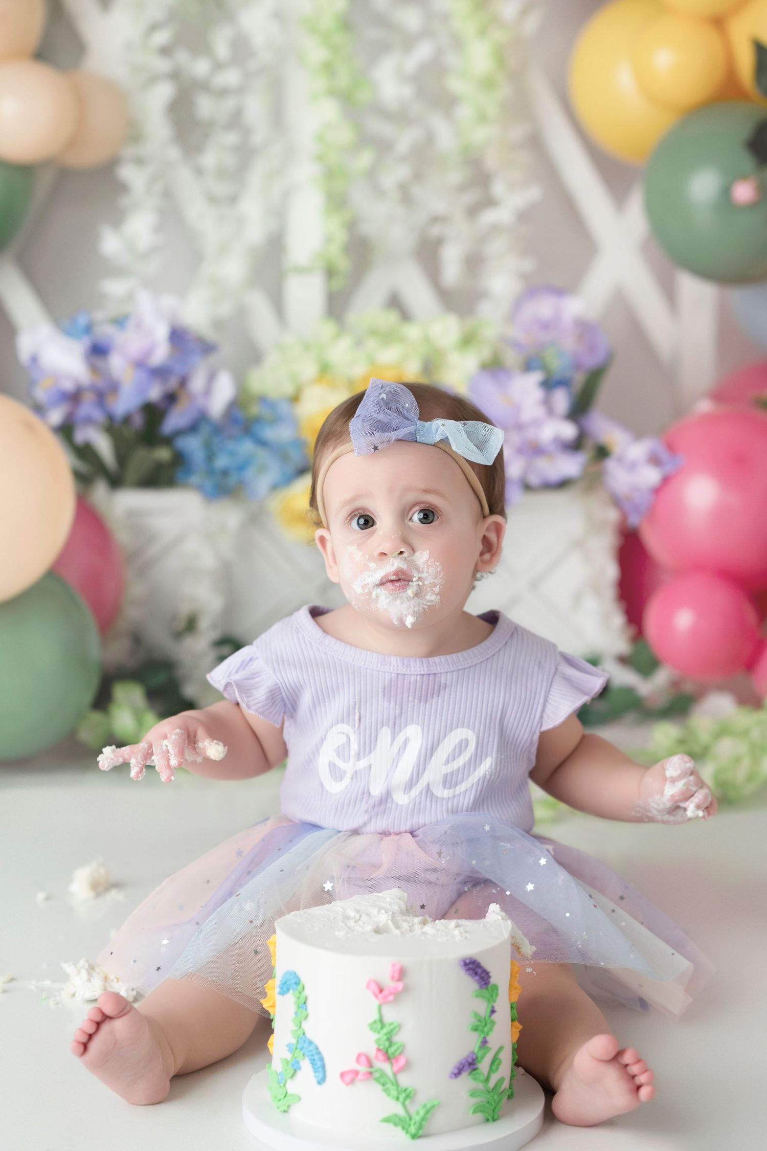 Baby girl in purple tutu celebrates first birthday smashing cake, with frosting on face, surrounded by colorful balloons.