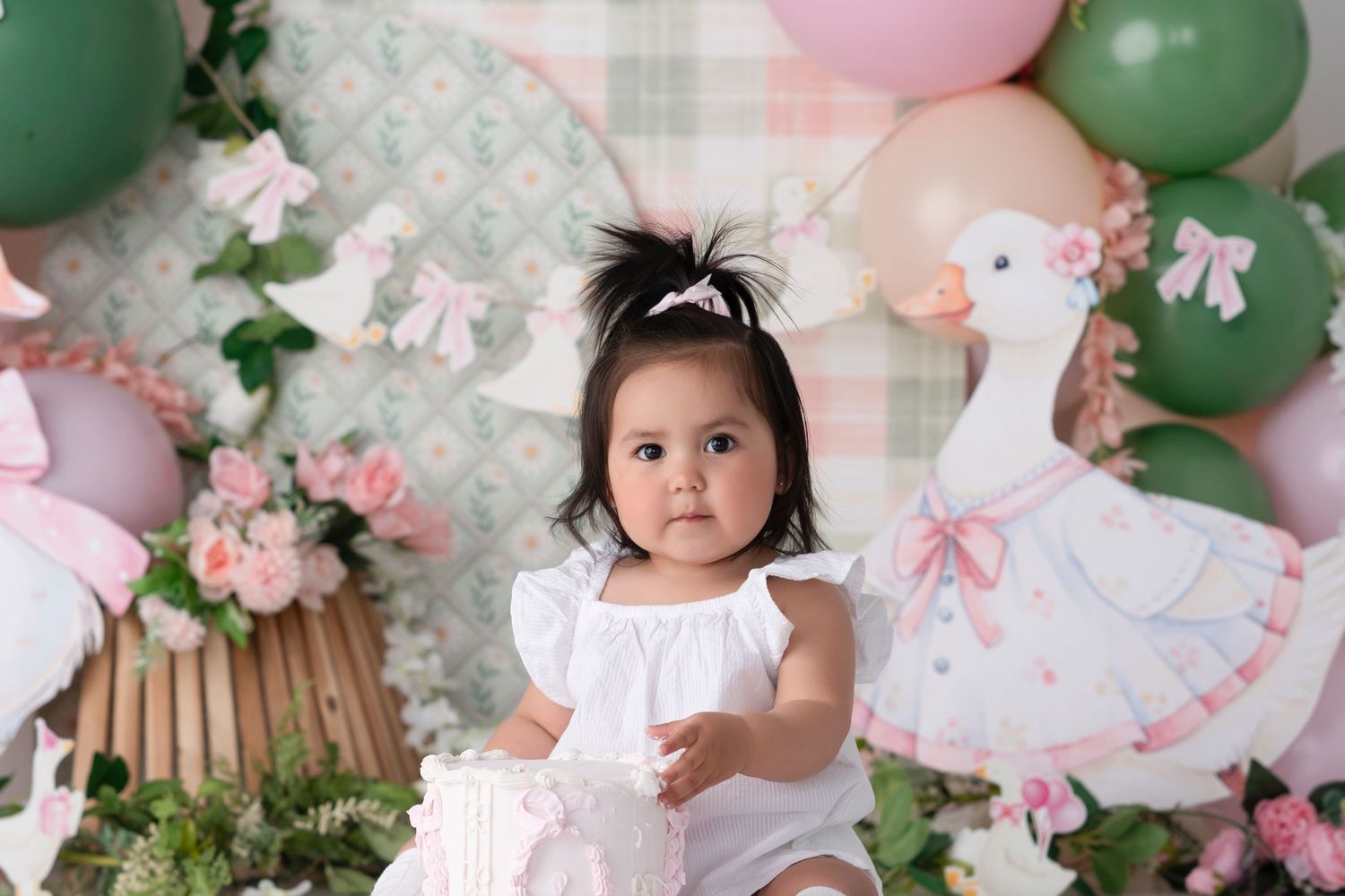 Baby girl in white dress sits by a cake with swan and balloon backdrop at a first birthday photoshoot.