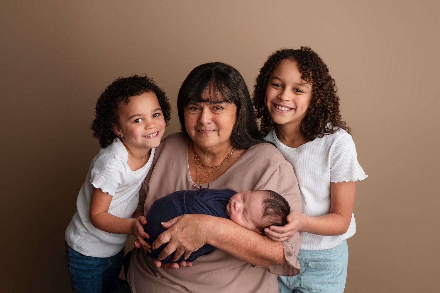 Grandmother holds newborn baby while two young girls smile beside her in a warm studio portrait.