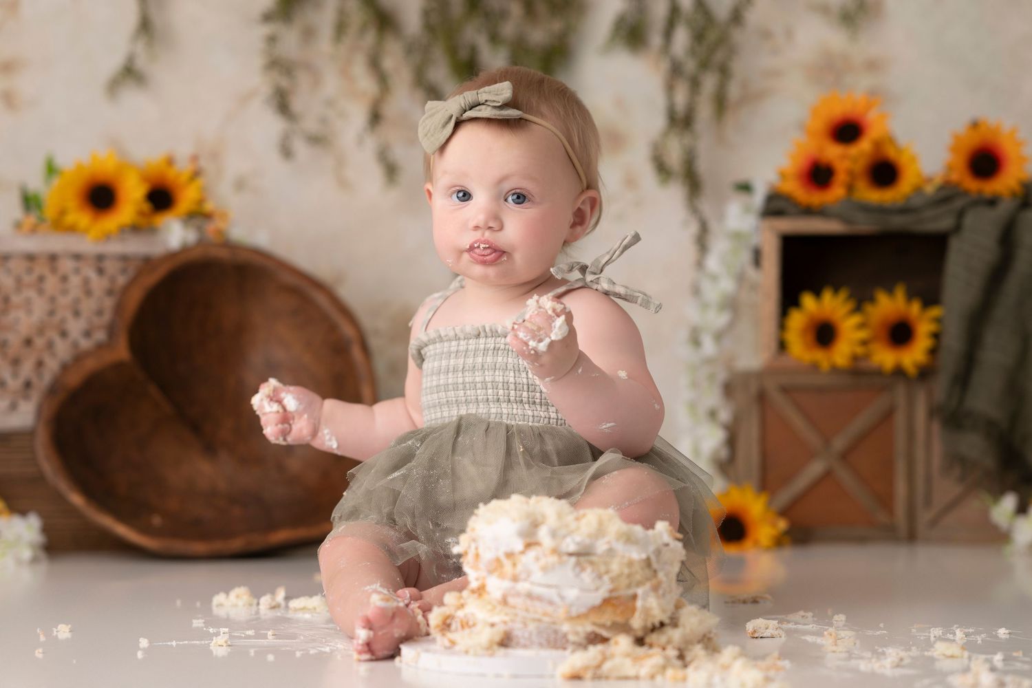 Baby girl in olive romper enjoys first birthday cake smash surrounded by sunflowers and rustic decor.