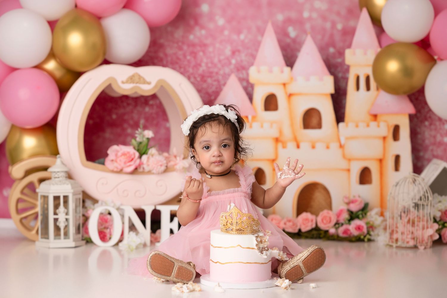 Baby girl in pink dress celebrates first birthday with castle backdrop, balloons, and smash cake.
