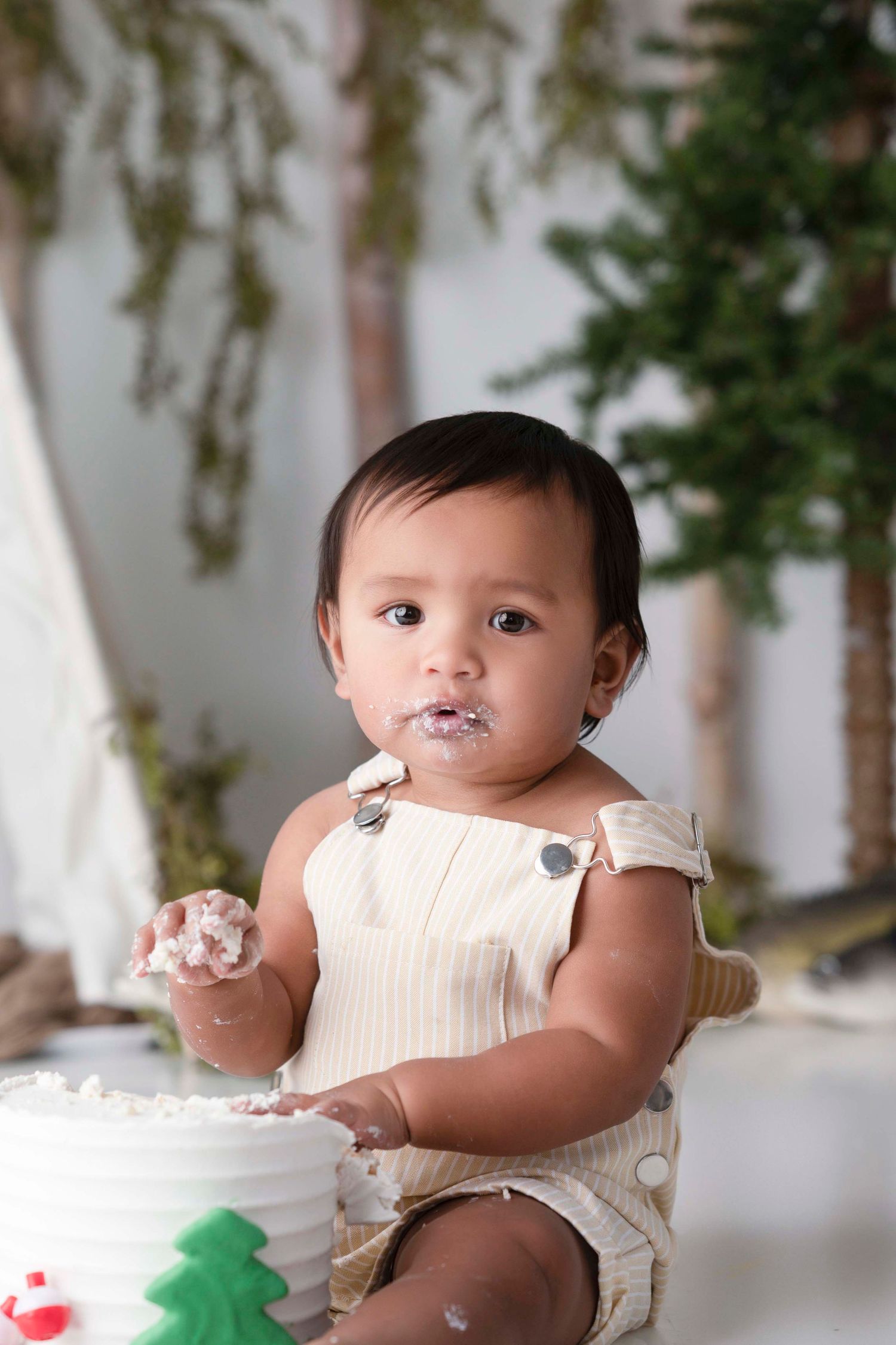 Cute baby girl smashing her first birthday cake, covered in white frosting, wearing a beige romper.