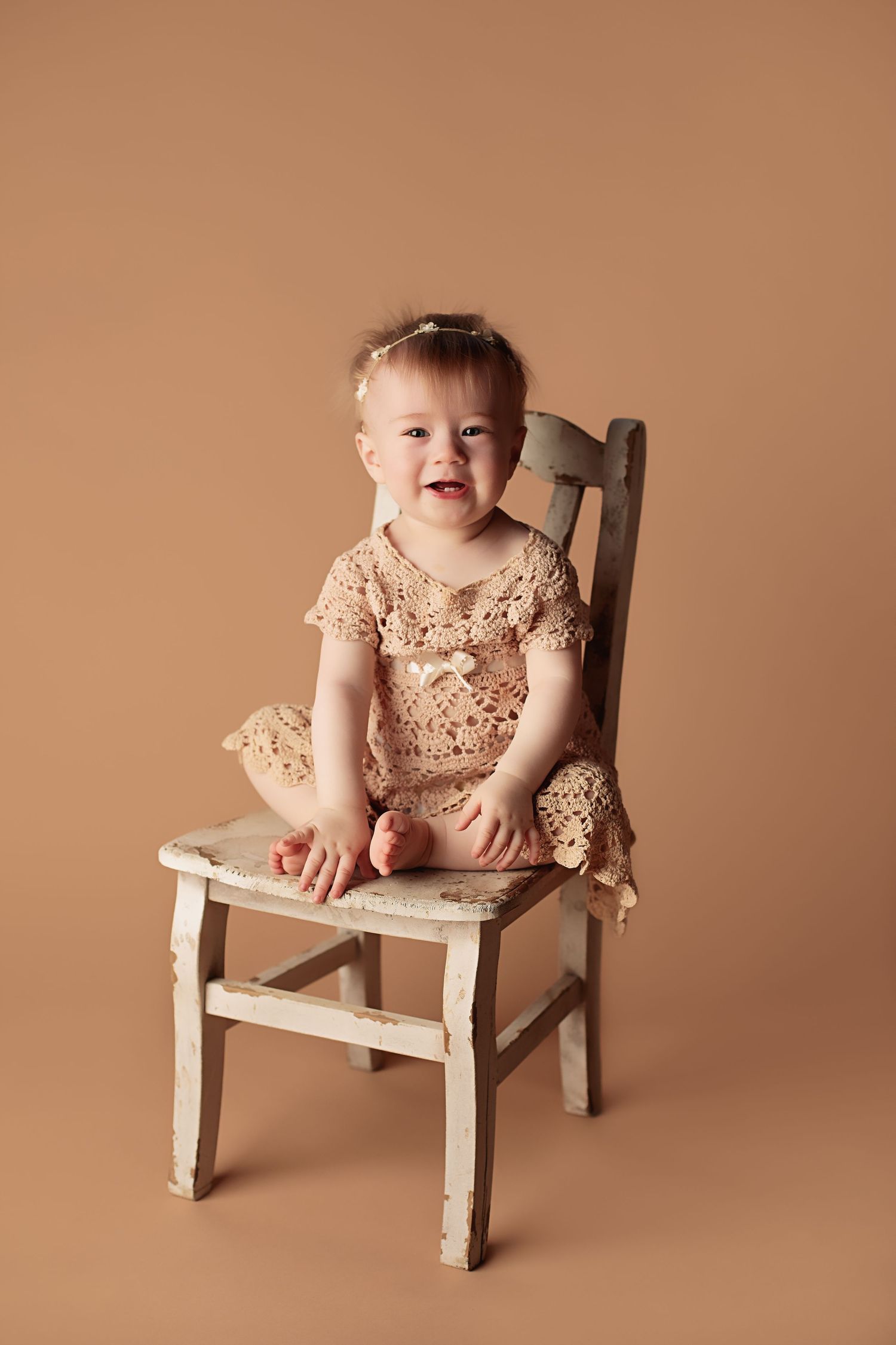 A toddler wearing a sparkly beige dress sits on a distressed white wooden chair against a brown backdrop.