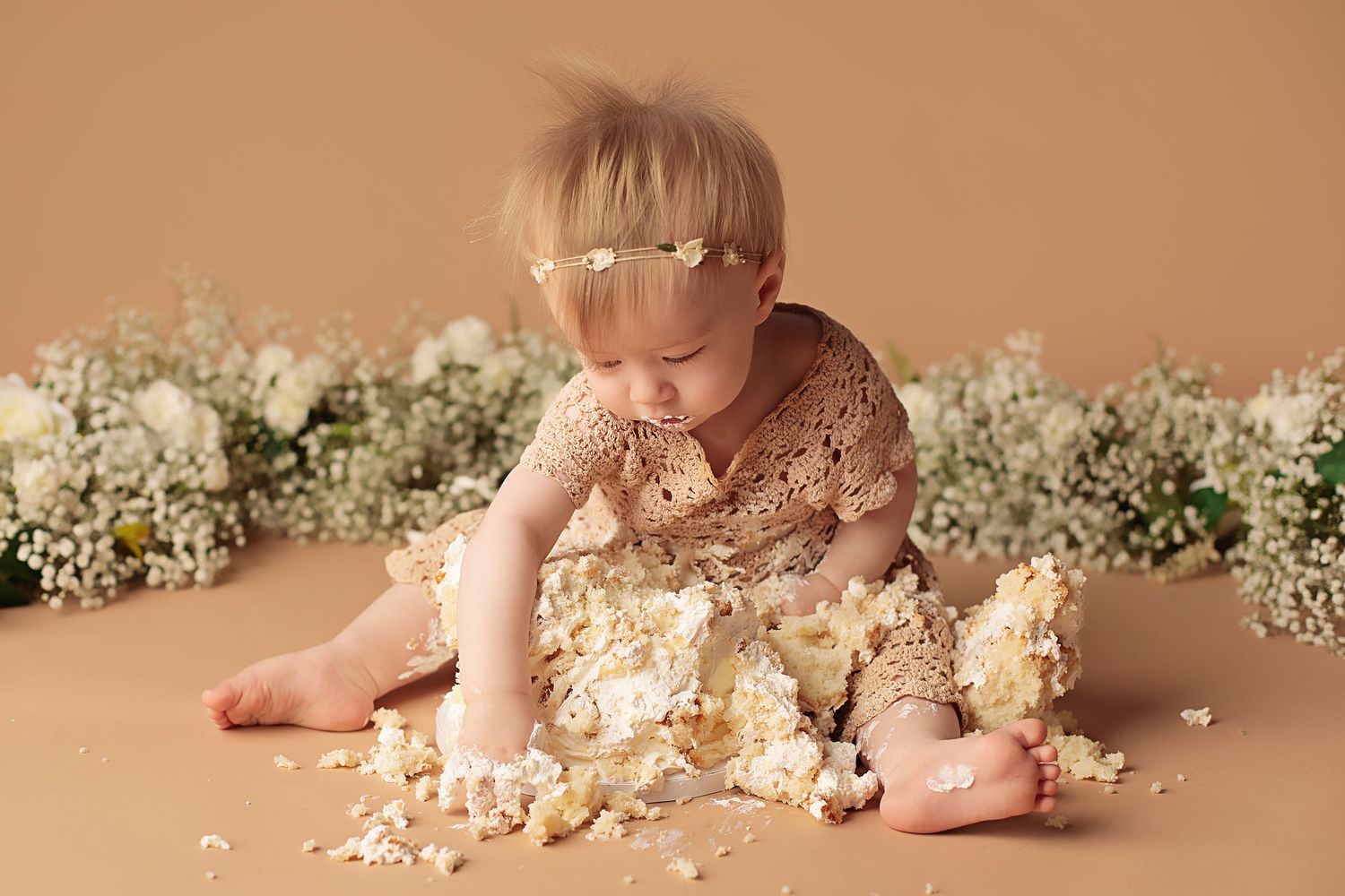 A baby in beige backdrop lies among white flowers while playing with cake in a first birthday cake smash photo session.