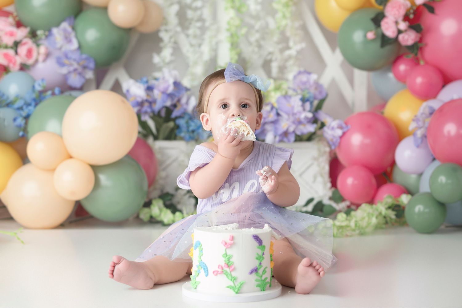 Baby sitting on floor with colorful balloon arch and floral decorations for milestone photo session.