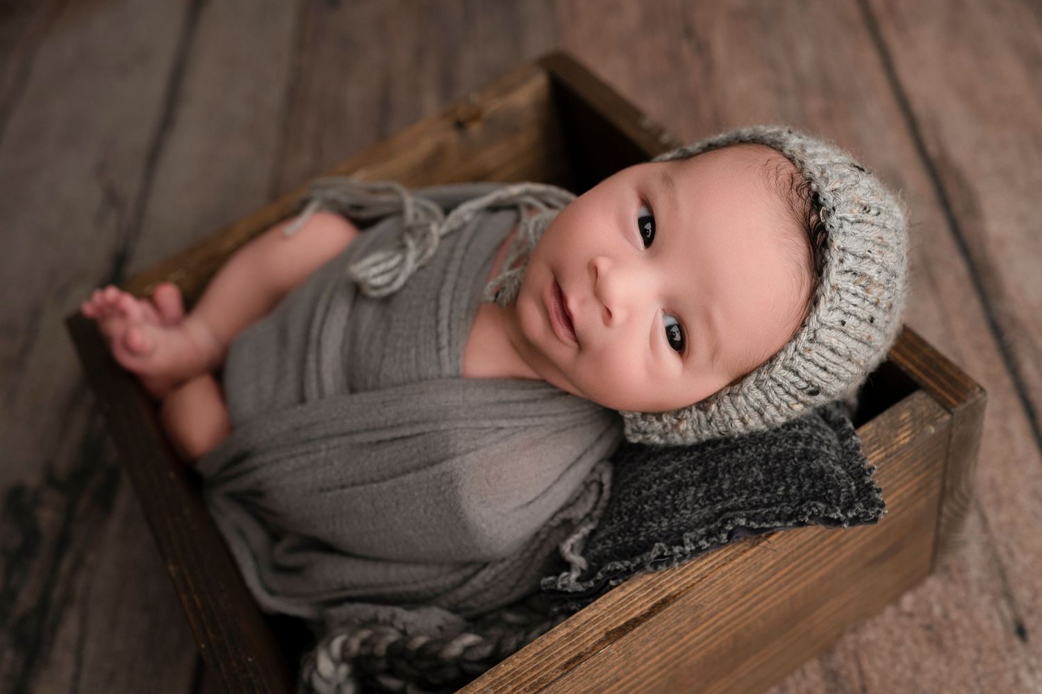 Newborn baby wrapped in gray swaddle wearing knit bonnet, resting in rustic wooden crate on wood floor.