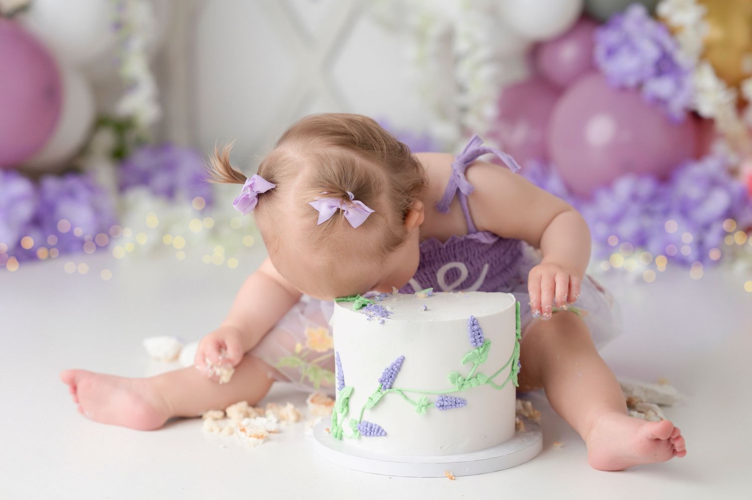 Baby girl in purple romper smashing her first birthday cake surrounded by purple balloons and floral decorations.